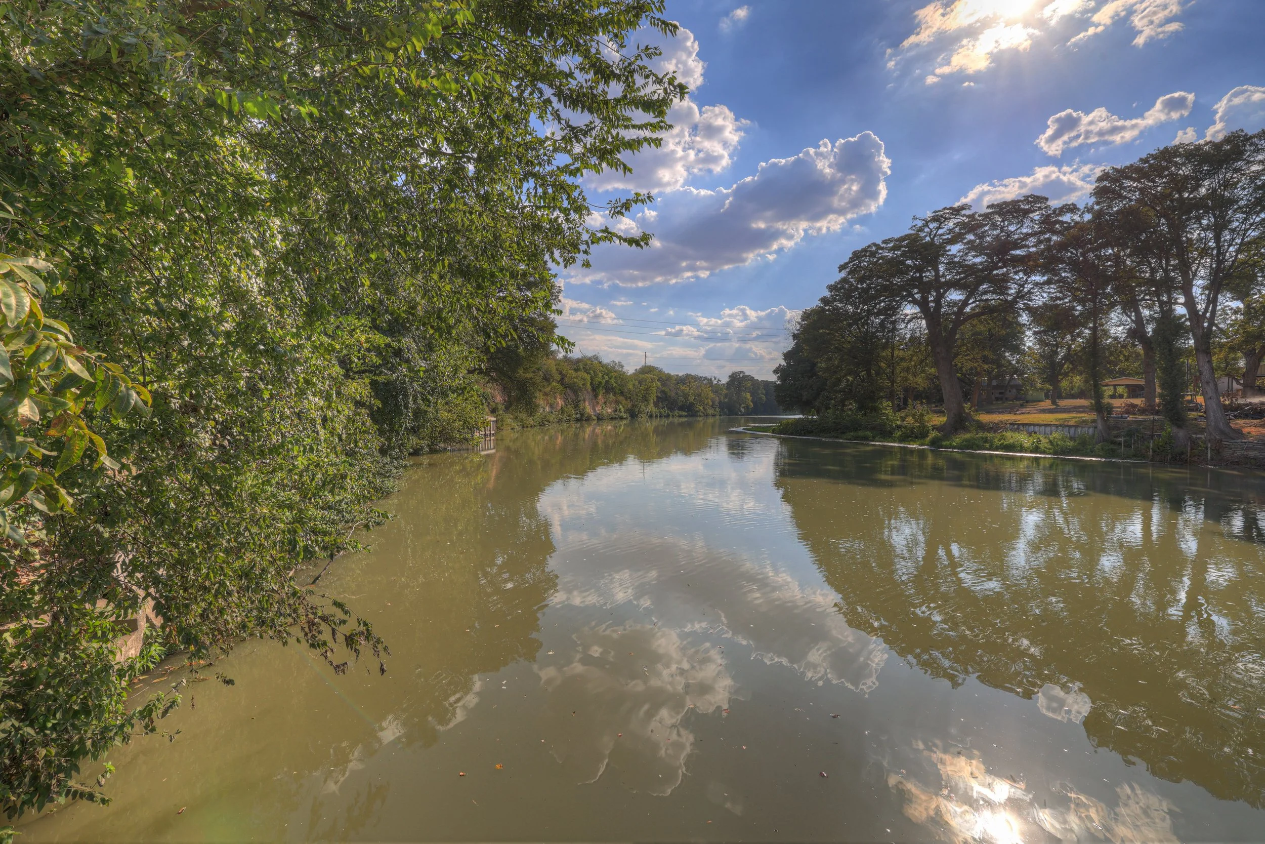 A peaceful river scene with calm water reflecting the blue sky and fluffy clouds, bordered by lush green trees on the left and taller trees on the right, with a few houses and a fence visible in the background.