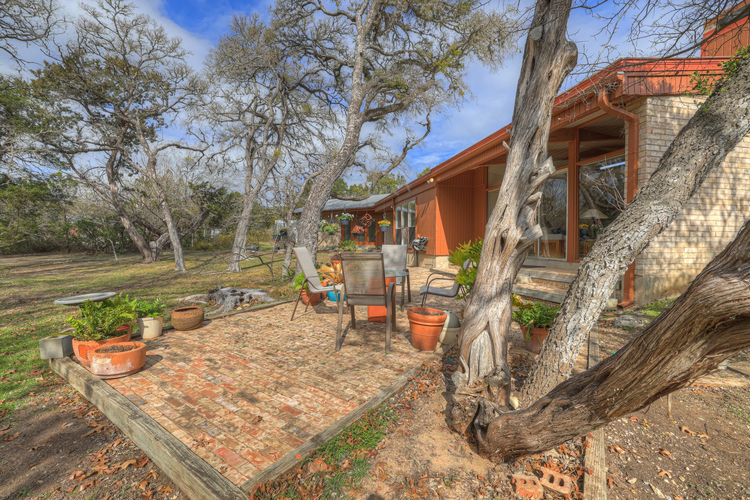 A backyard patio with brick flooring, surrounded by trees and potted plants, with outdoor chairs and a table, adjacent to a house with large windows and brick and wood exterior.
