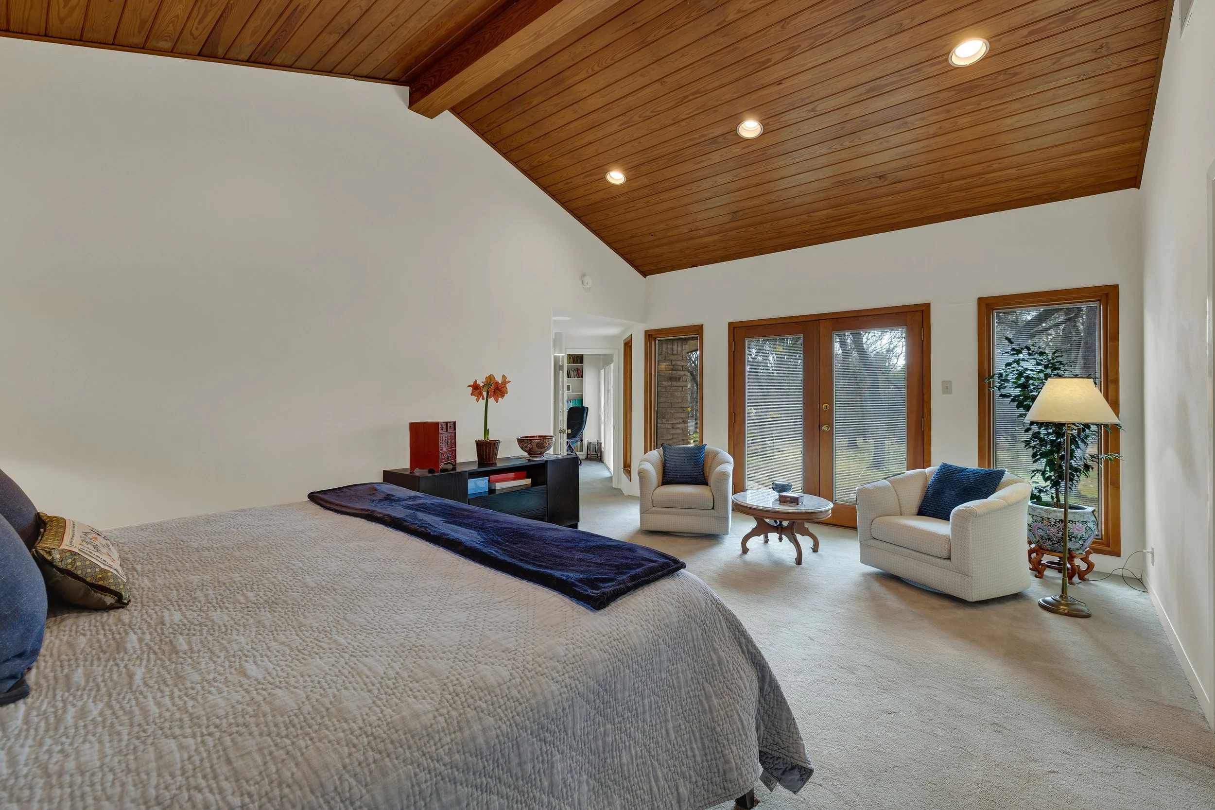 A spacious bedroom with a sloped wooden ceiling, white walls, and large windows. The room contains a bed with a beige quilt, two white armchairs with pillows, a round wooden table, a floor lamp, and a black dresser against the wall. There are double 