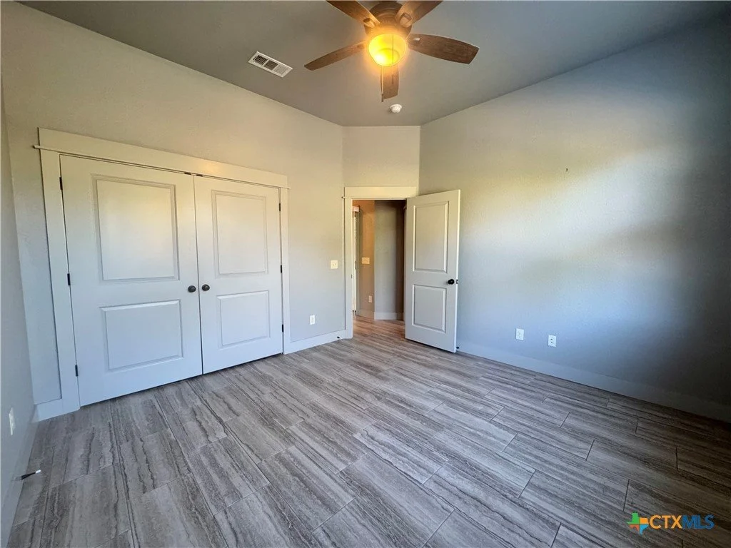 Empty bedroom with light gray walls, wood-look tile flooring, a ceiling fan, a partially open door, and a closed closet door.