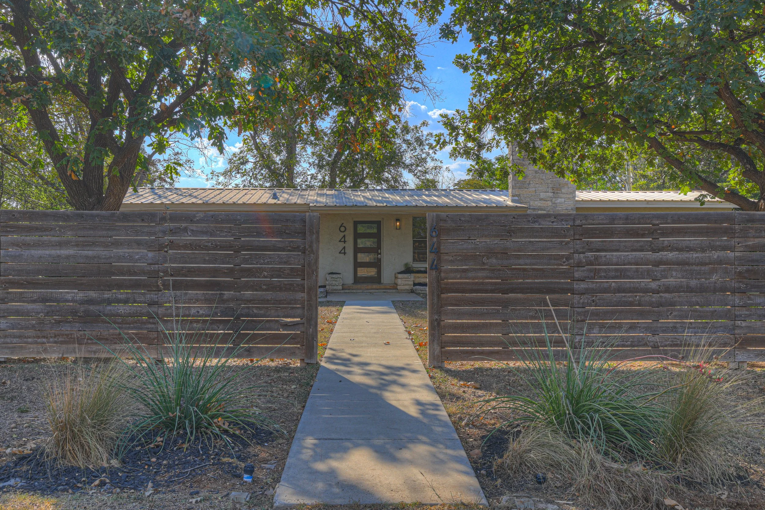 Front view of a house enclosed by a tall wooden fence, with a concrete walkway leading to the front door, surrounded by plants and trees under a partly cloudy sky.