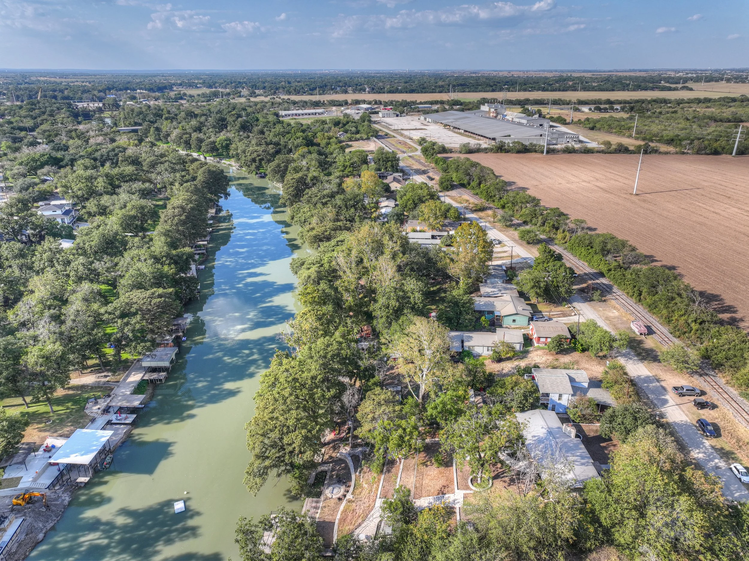Aerial view of a canal lined with trees and houses with boat docks on the left side, and a rural area with fields, trees, and a row of small buildings on the right side, under a partly cloudy sky.