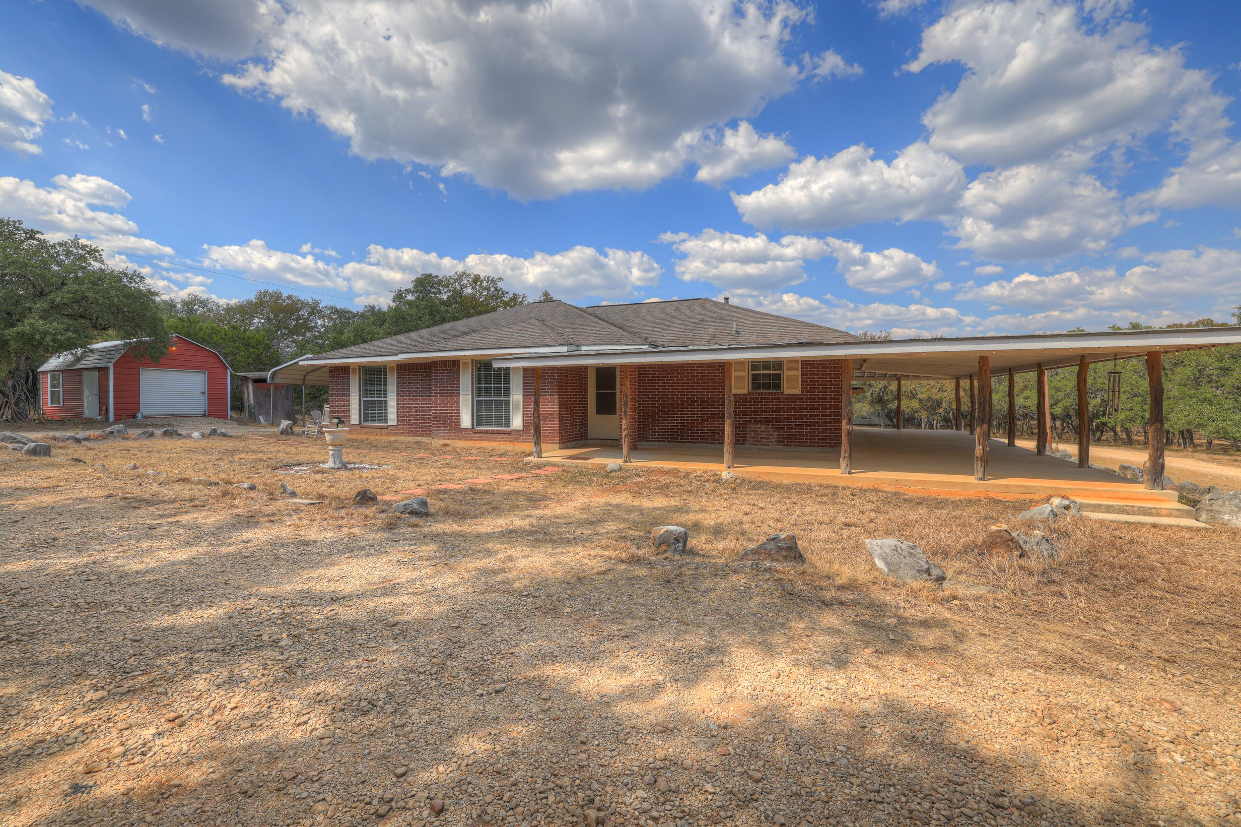A brick house with a covered porch and a gravel yard under a partly cloudy sky, with a small red shed in the background.