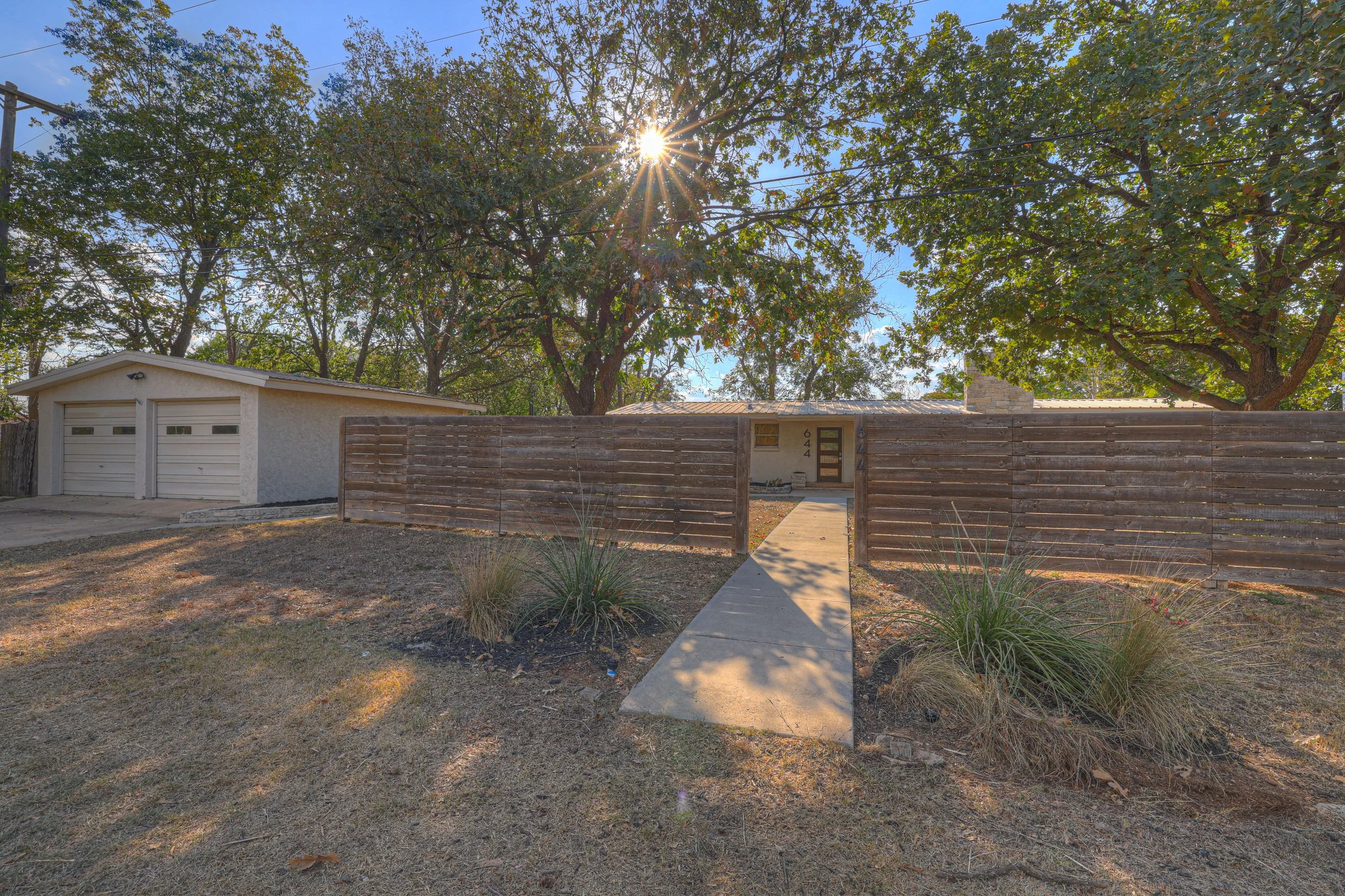 Front view of a house with a wooden fence, sidewalk leading to the door, and a detached garage, surrounded by trees, with sunlight shining through the leaves.