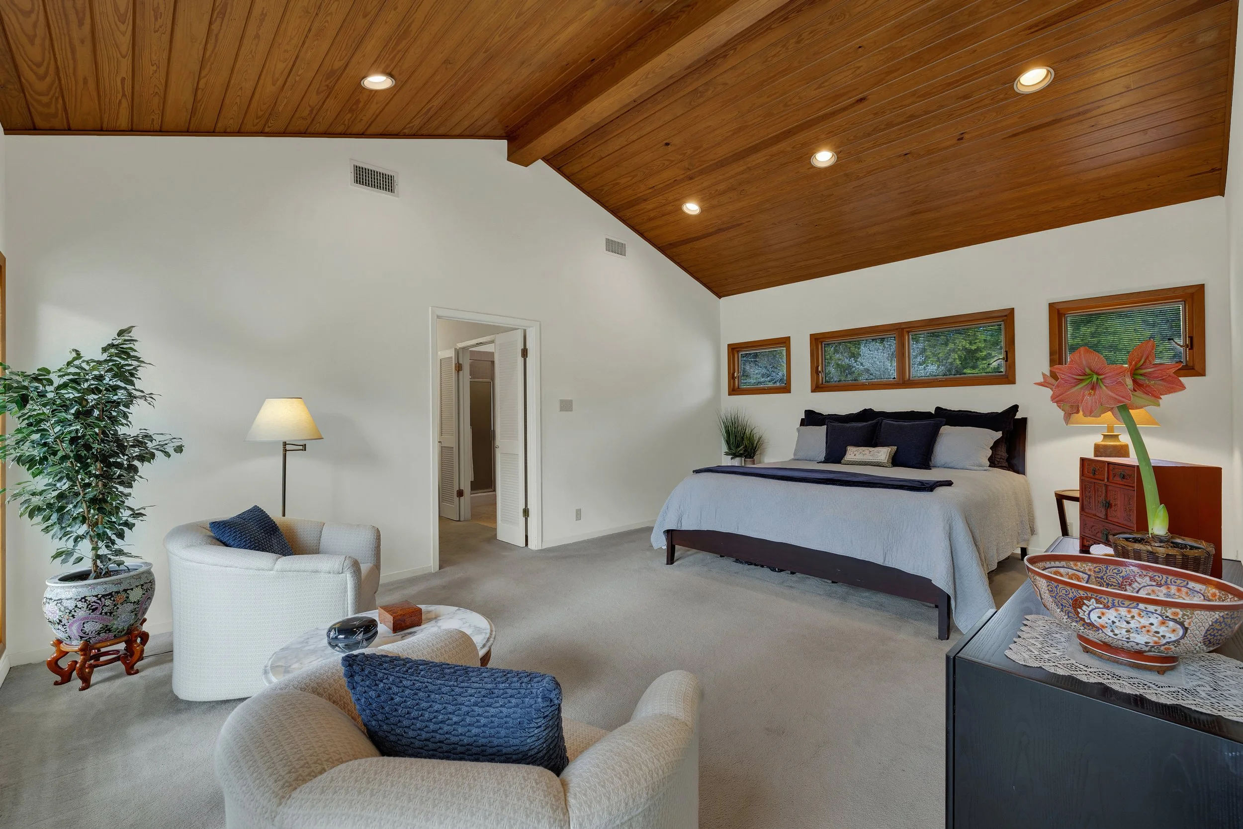A bedroom with a wooden ceiling, white walls, and neutral carpet. There is a large bed with multiple pillows, a small nightstand, and three small horizontal windows above the headboard. To the left, there is a sitting area with two chairs, a small ta