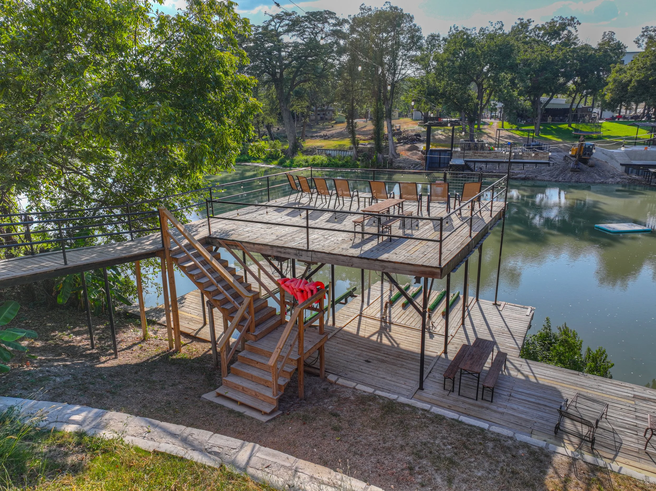 A wooden deck with outdoor chairs and tables is built over a body of water, with stairs leading up to it and railings along the edges, surrounded by trees.