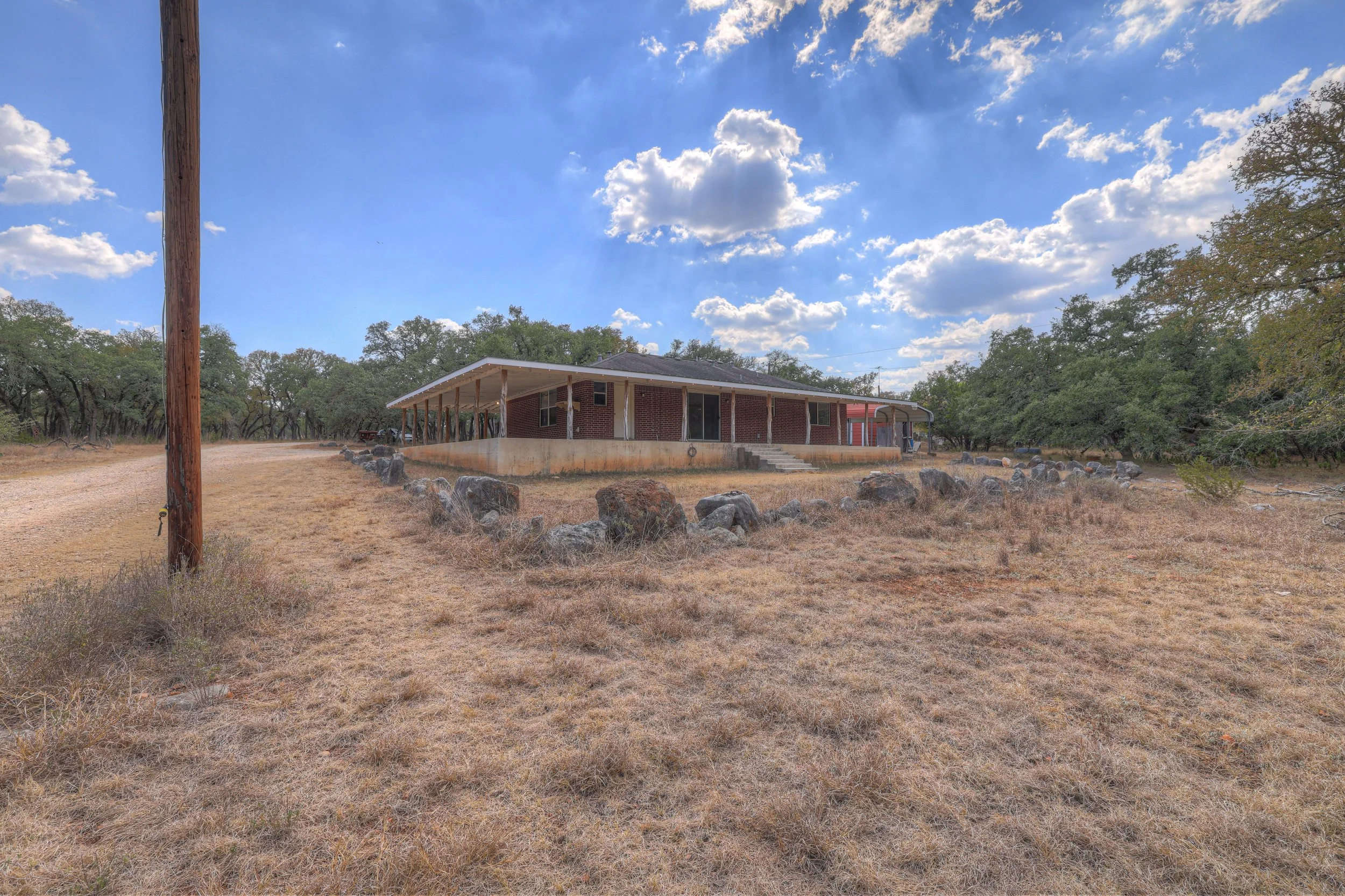 A single-story brick house with a porch, set in a dry landscape with sparse grass, surrounded by a rocky border and trees under a partly cloudy sky.