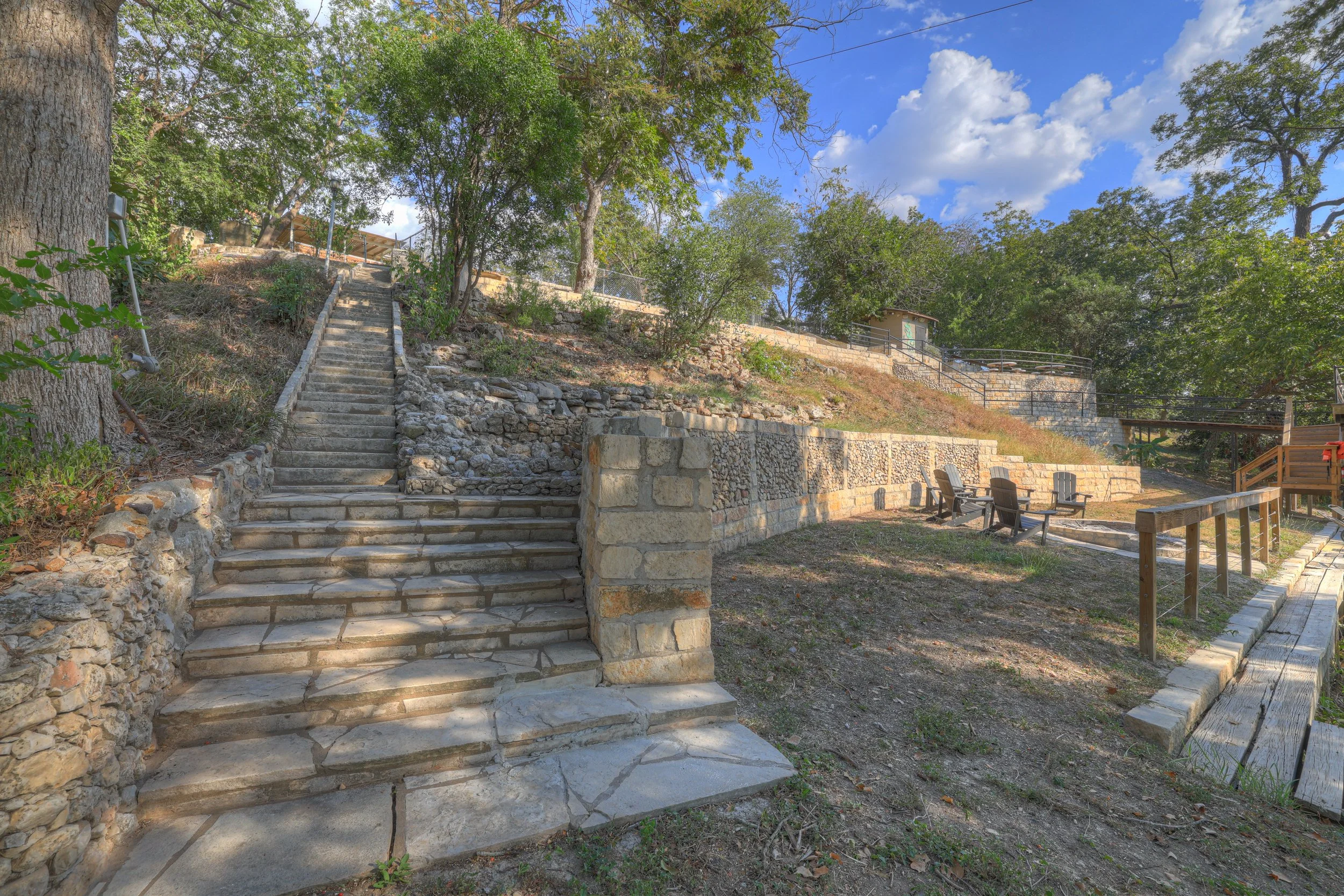Stone stairway leading up a hillside, surrounded by trees and stone walls, with a small seating area and wooden fencing nearby, under a partly cloudy sky.