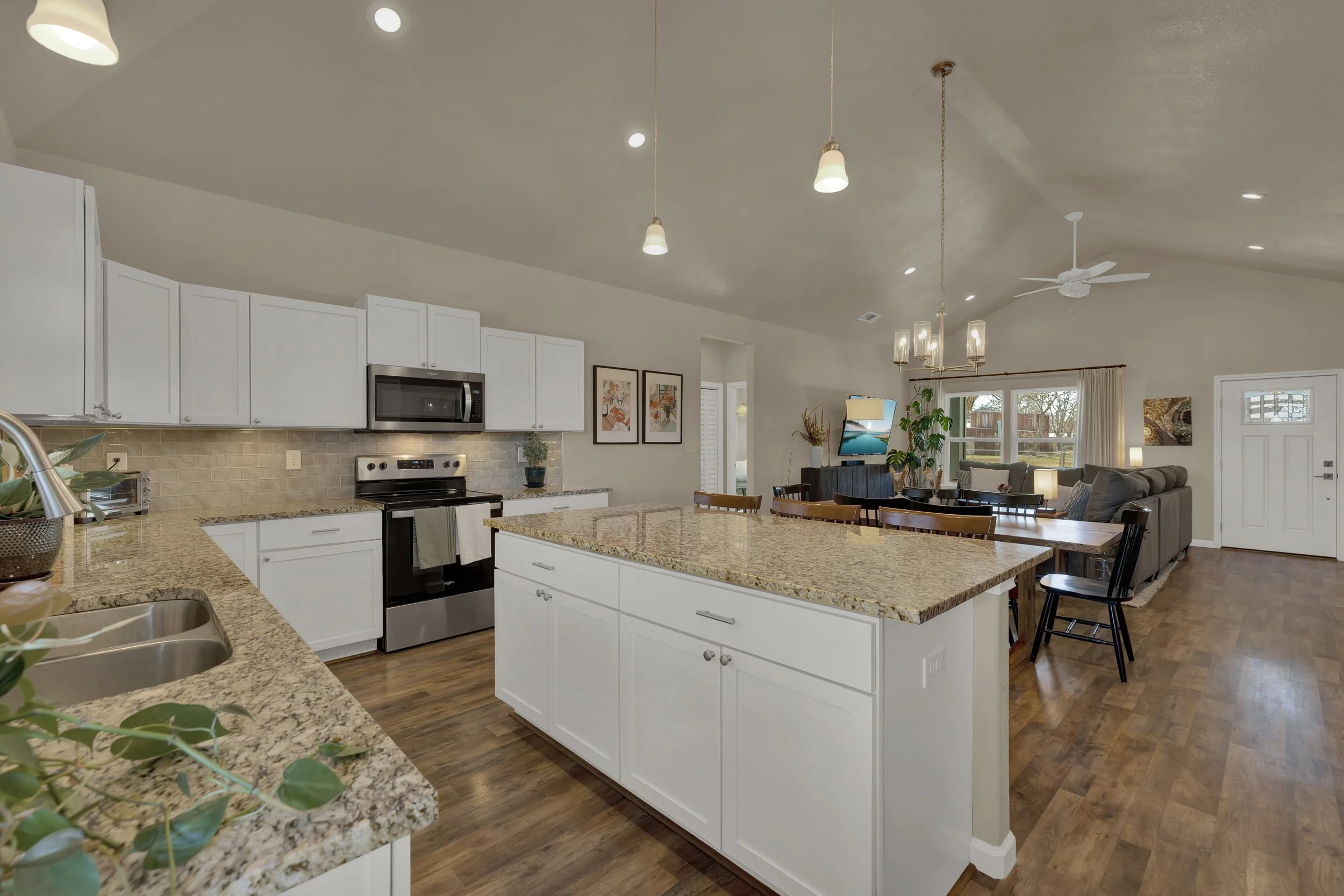 Spacious open-concept kitchen and living room with white cabinets, granite countertops, stainless steel appliances, hardwood floors, overlapping kitchen and living areas, pendant and chandelier lighting, and large windows with curtains.