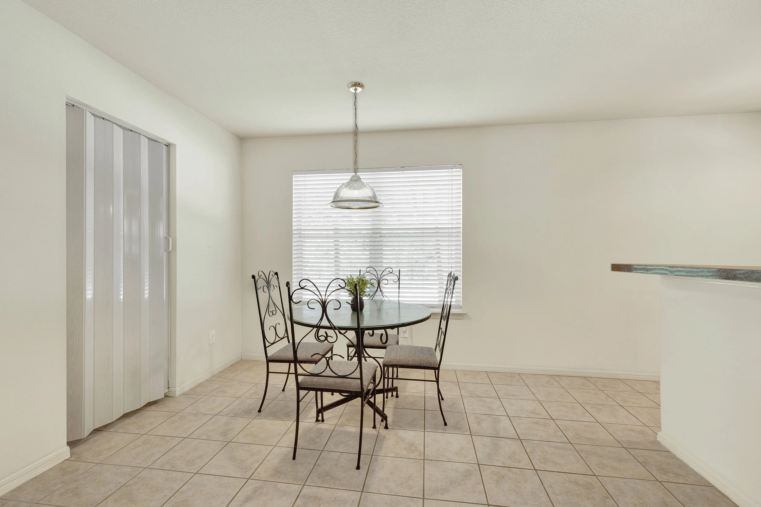 Empty dining area with a round table, four metal chairs with cushioned seats, a window with blinds, tiled floor, and a hanging light fixture.