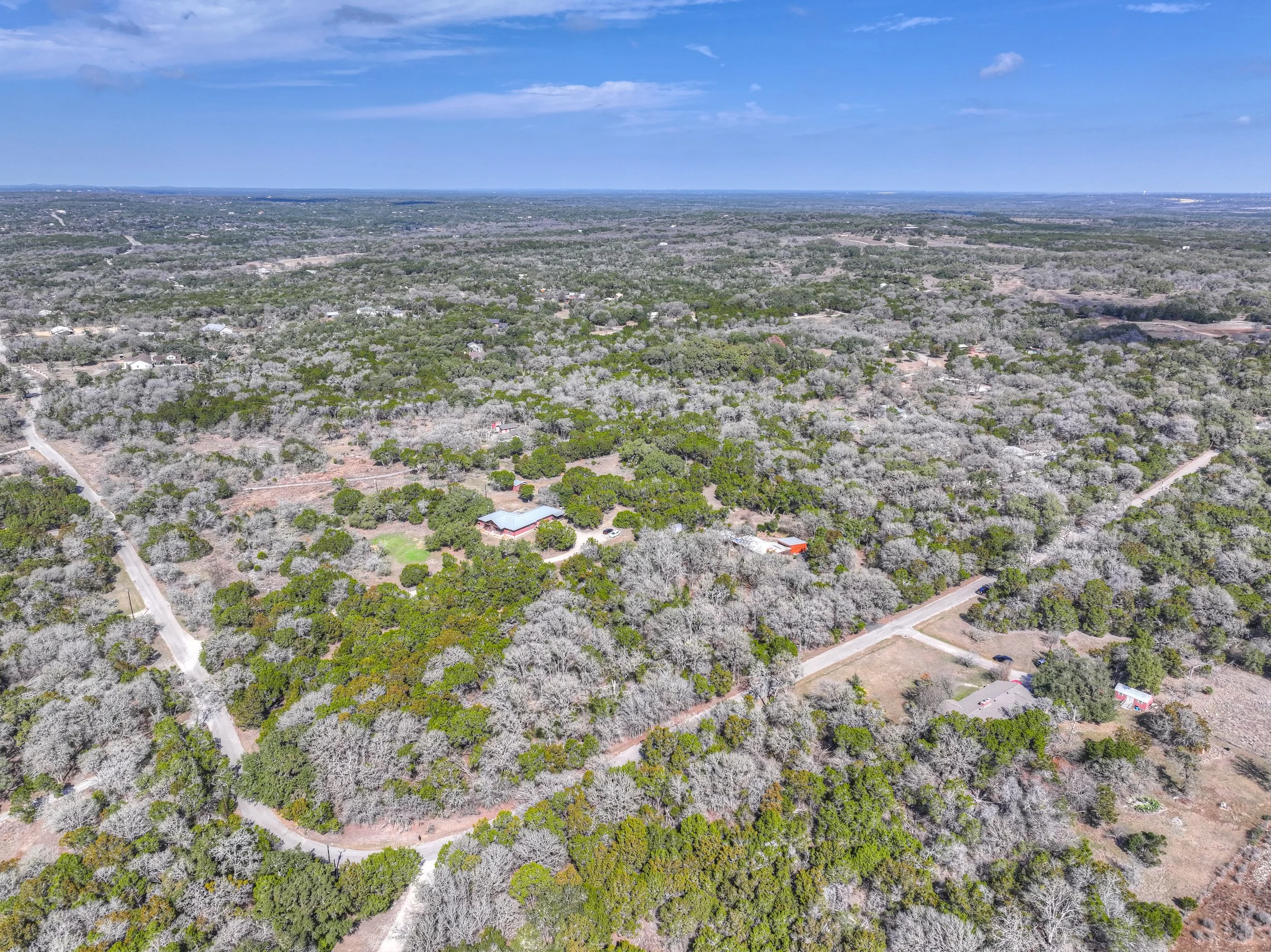 Aerial view of a rural landscape with scattered trees, dirt roads, and a few houses under a blue sky with some clouds.
