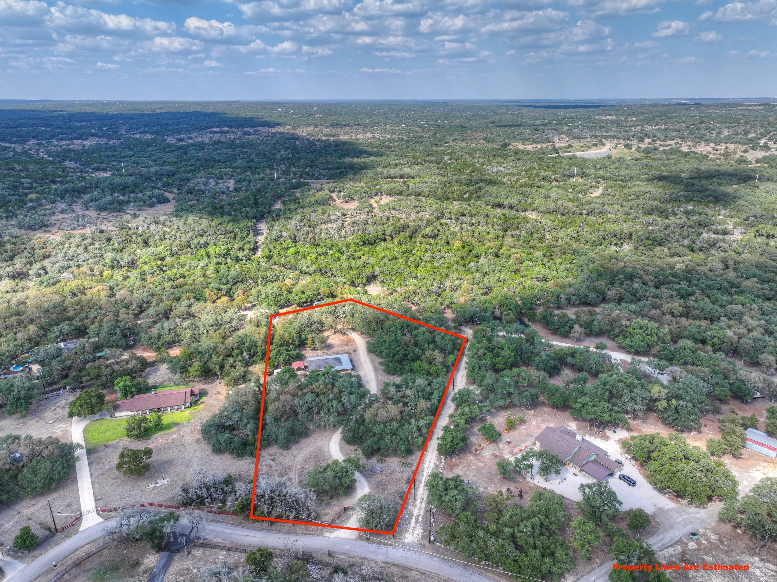 Aerial view of a rural area with a red property line outlining a wooded lot with a driveway and a building, surrounded by other houses and dense trees, under a blue sky with some clouds.