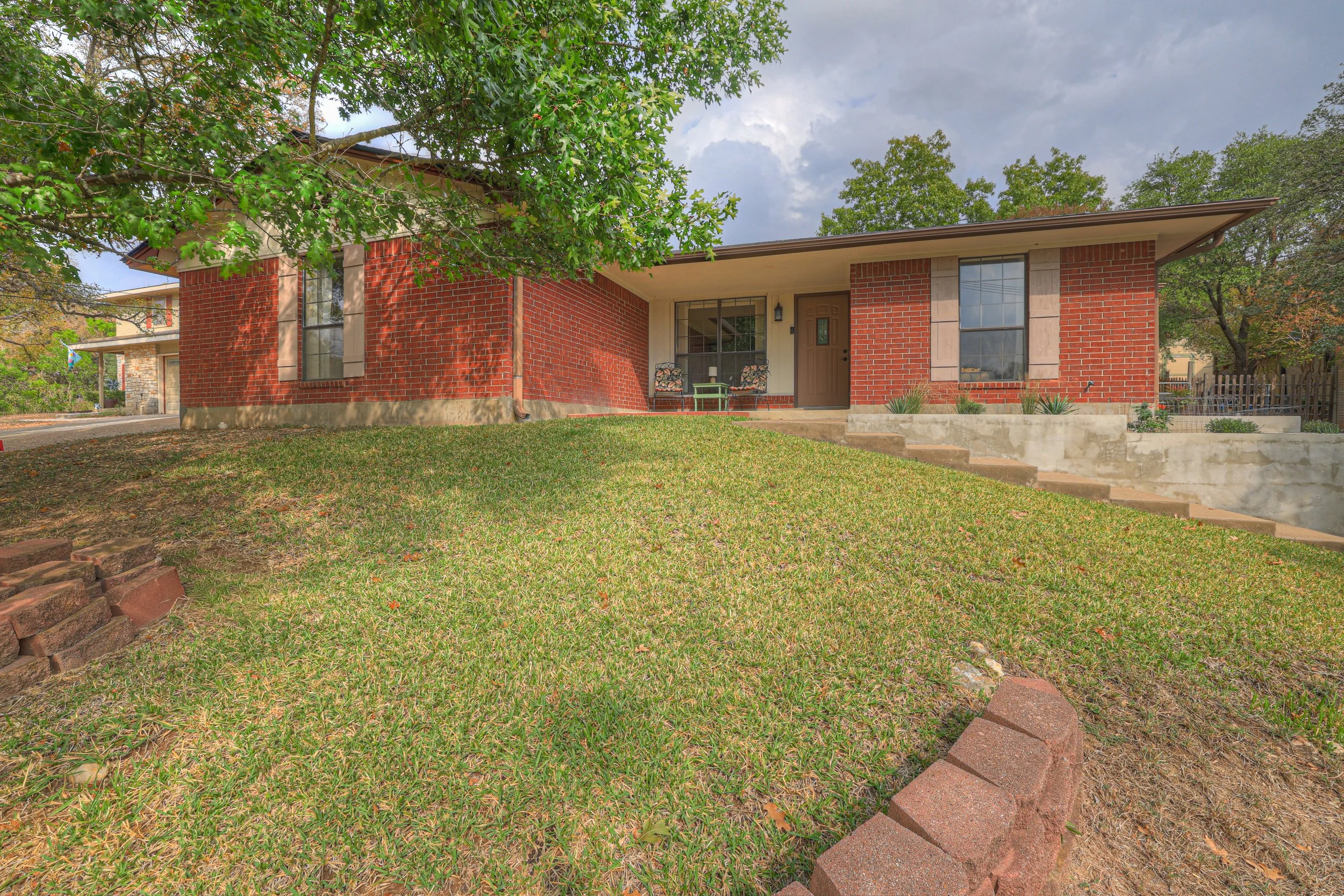 A red brick house with a porch, two chairs, and a small table on the front porch, surrounded by a green lawn and trees, under a partly cloudy sky.
