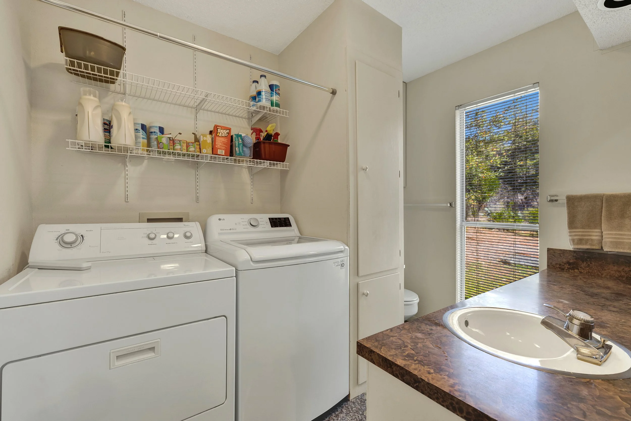 Laundry room with a top-loading washing machine and a front-loading dryer, shelves with laundry supplies above, a window with blinds, a countertop with a sink, and a small toilet behind a cabinet.