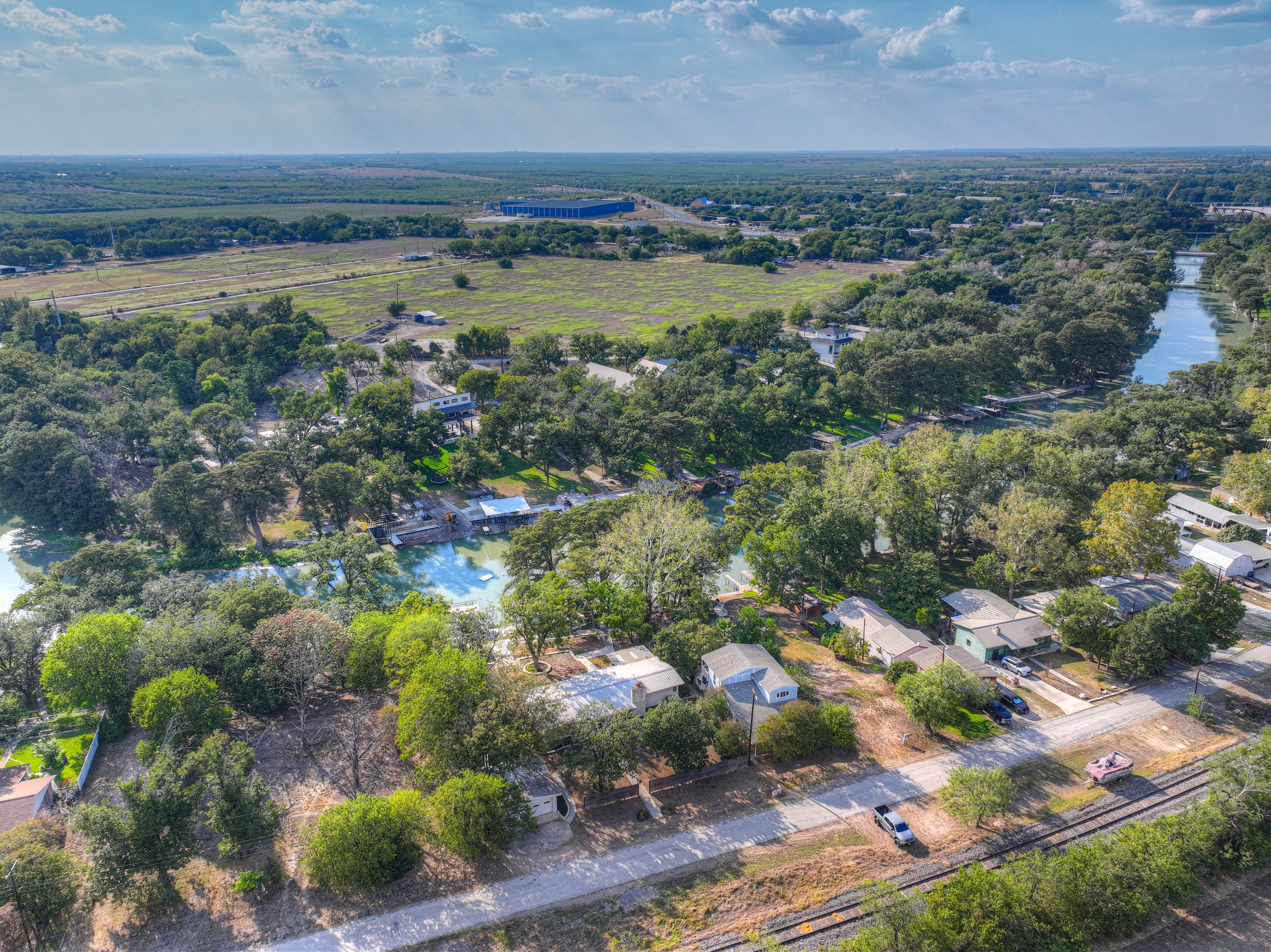 Aerial view of a neighborhood with houses, trees, a river, and open fields under a partly cloudy sky.