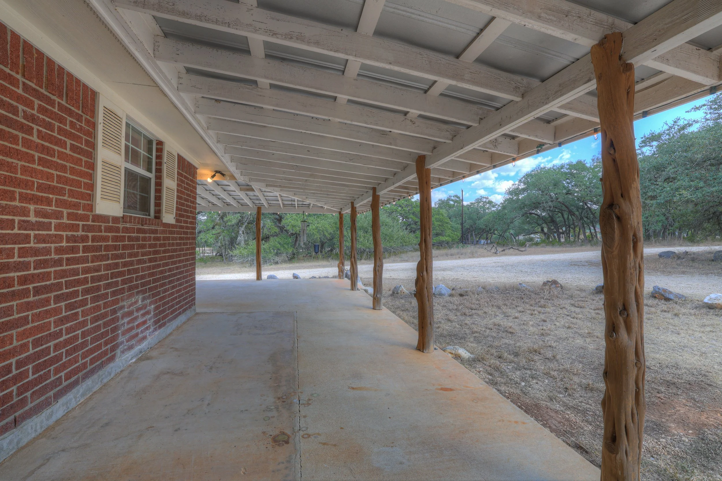 A covered patio area with a concrete floor and wooden support beams outside a brick house, overlooking a grassy yard with trees and rocks under a partly cloudy sky.
