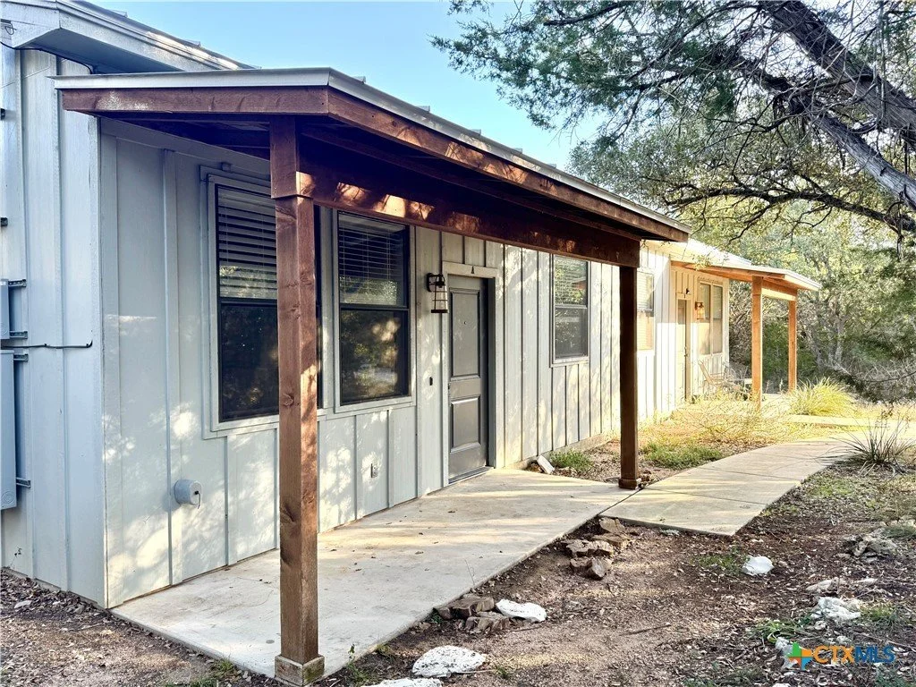 Exterior view of a house with two porches, one darker wood and one lighter, with a walkway leading to the front door, surrounded by trees and yard.