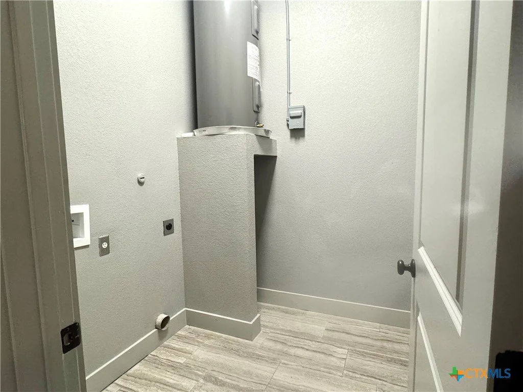 Empty laundry room with a water heater on a raised platform in the corner, electrical outlets, a vent pipe near the floor, and a door slightly open.