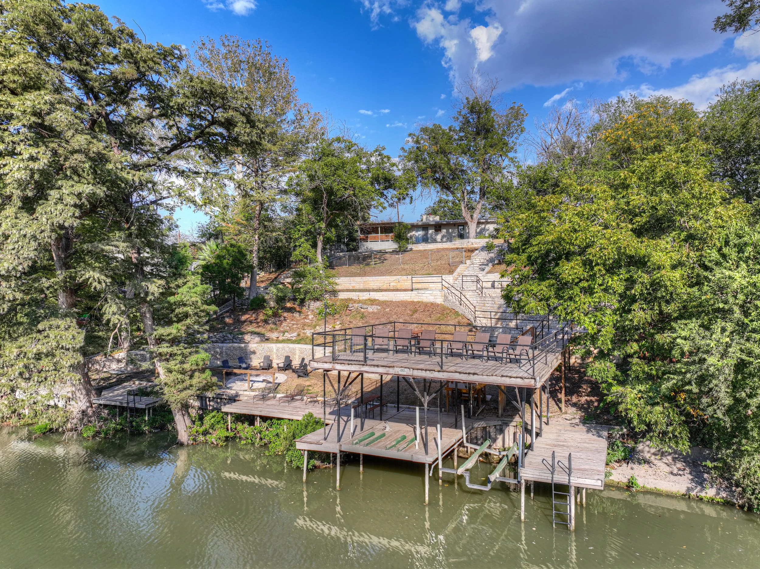 A multi-level wooden deck with railings extends over a canal, surrounded by trees and a hillside with stairs and different levels of outdoor seating, under a partly cloudy blue sky.