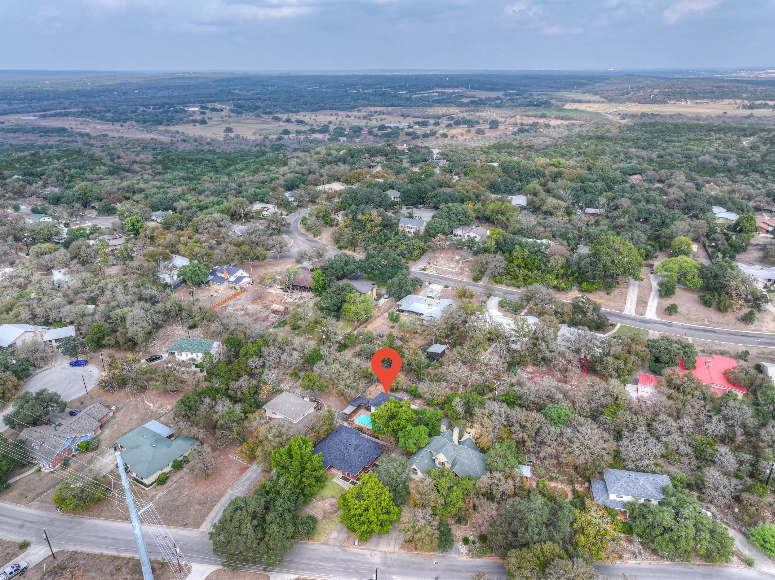 Aerial view of a suburban neighborhood with single-family houses, trees, and streets, with a red location marker pointing to a specific house.