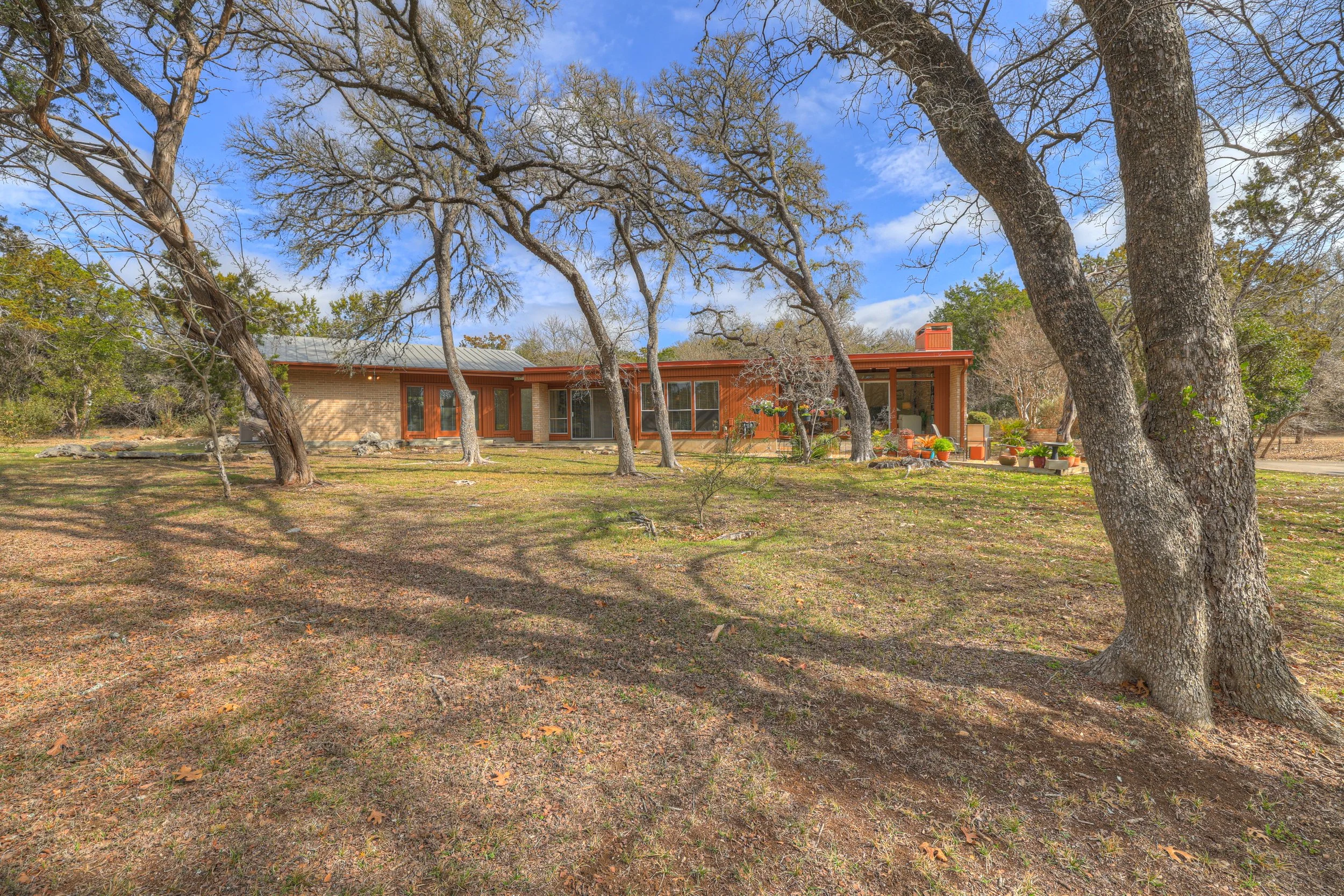 A mid-century modern house with a metal roof and extensive windows, surrounded by leafless trees and a grassy yard with shadows cast by the trees, under a partly cloudy sky.