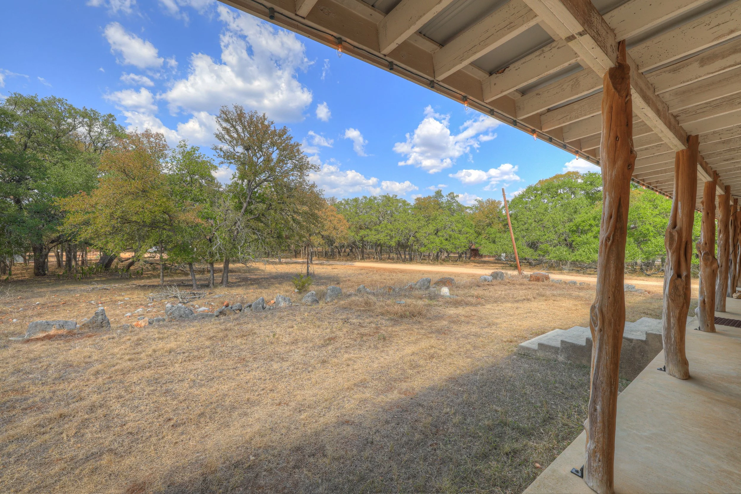 View from a covered porch with rustic wooden supports, overlooking a dry grassy field, trees with green and orange leaves, in a rural area under a blue sky with scattered clouds.