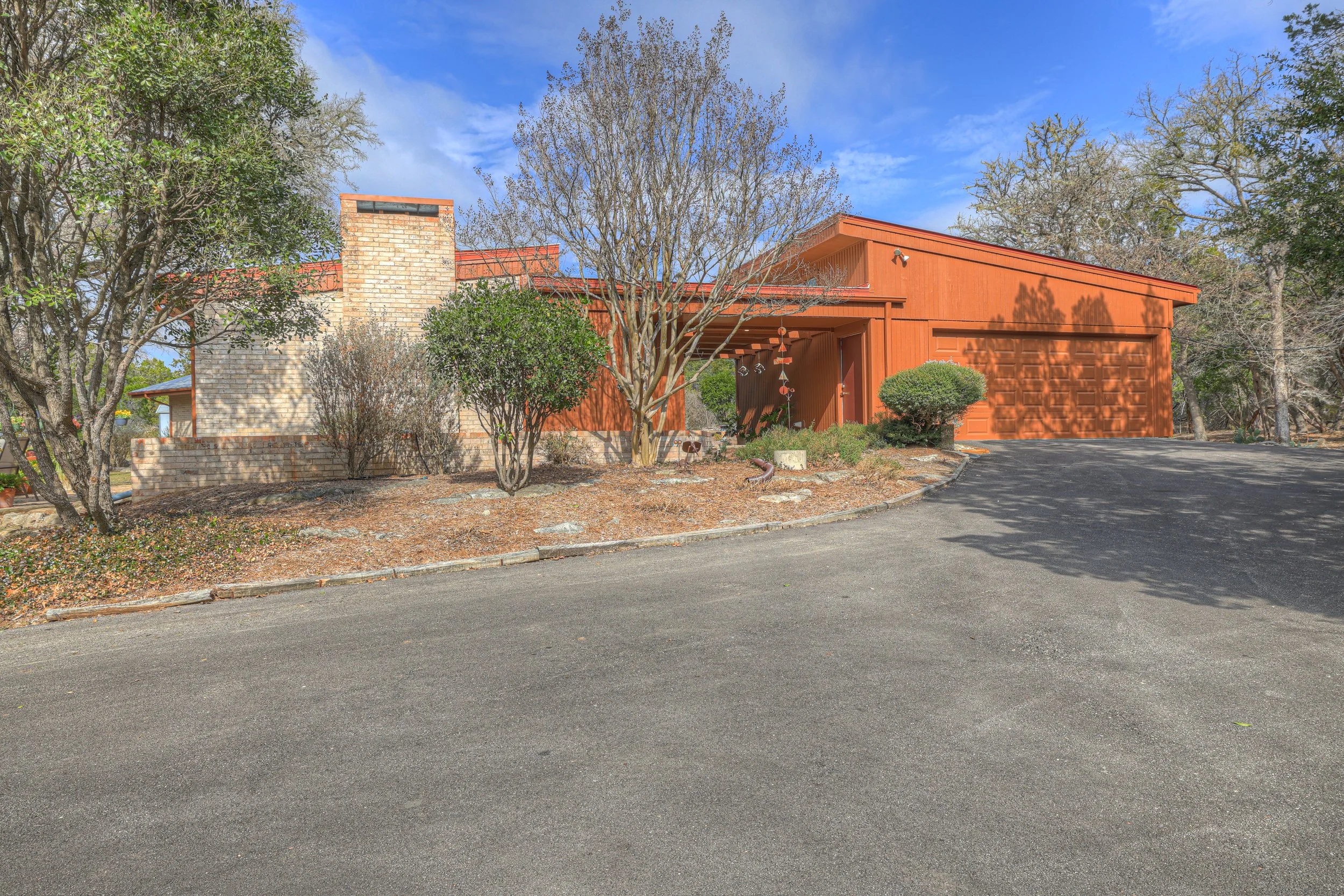 A modern house with a red-orange exterior, surrounded by trees and shrubs, with a driveway leading up to a closed garage door.
