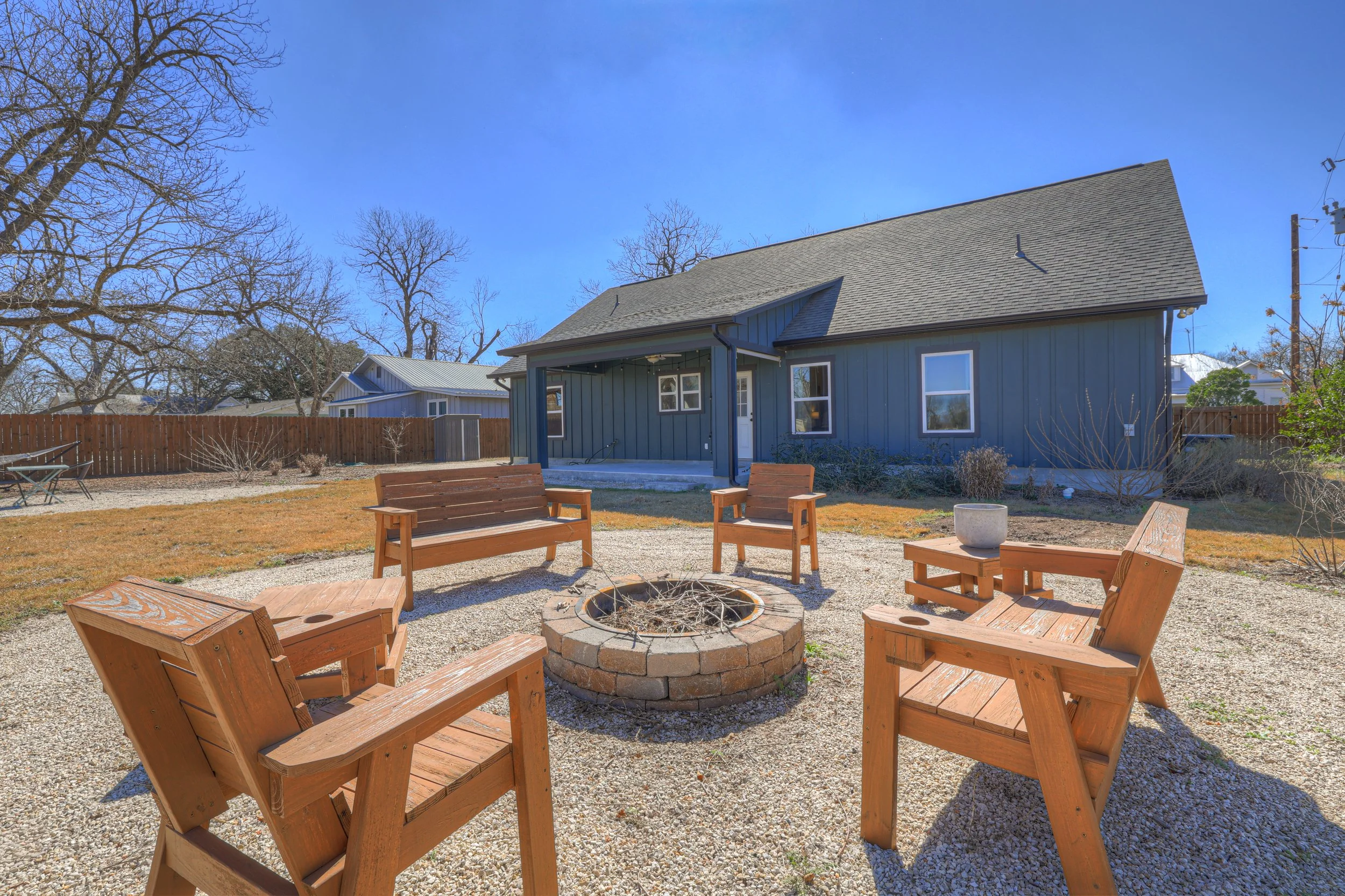 Backyard with wooden benches and chairs arranged around a fire pit, yellow grass, leafless trees, and a blue house with a covered porch under a clear blue sky.