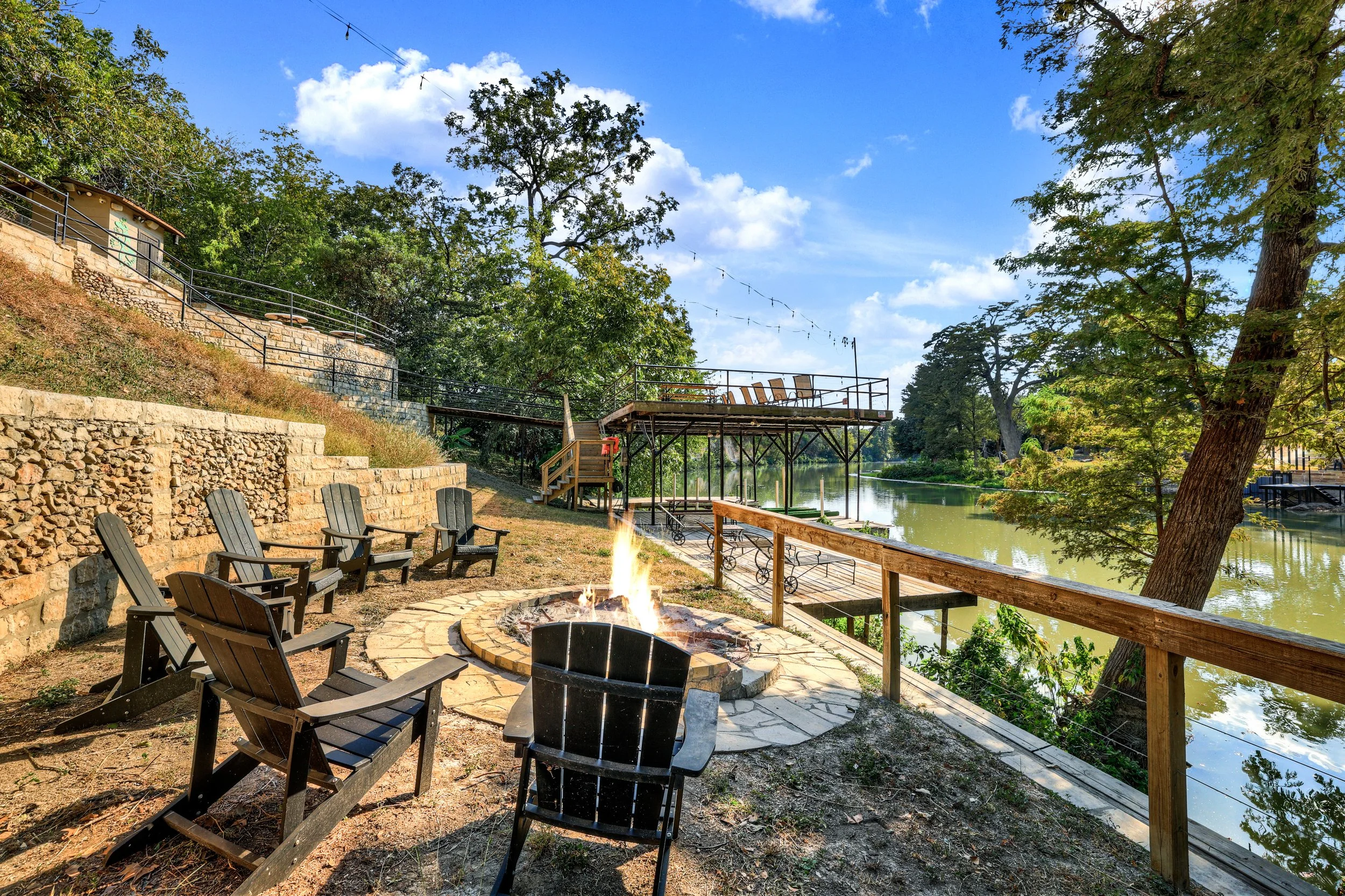 A lakeside outdoor patio with six black Adirondack chairs arranged around a fire pit with a small open flame. A stone pathway encircles the fire pit, and there is a railing along the water's edge. In the background, there is a raised deck with chairs