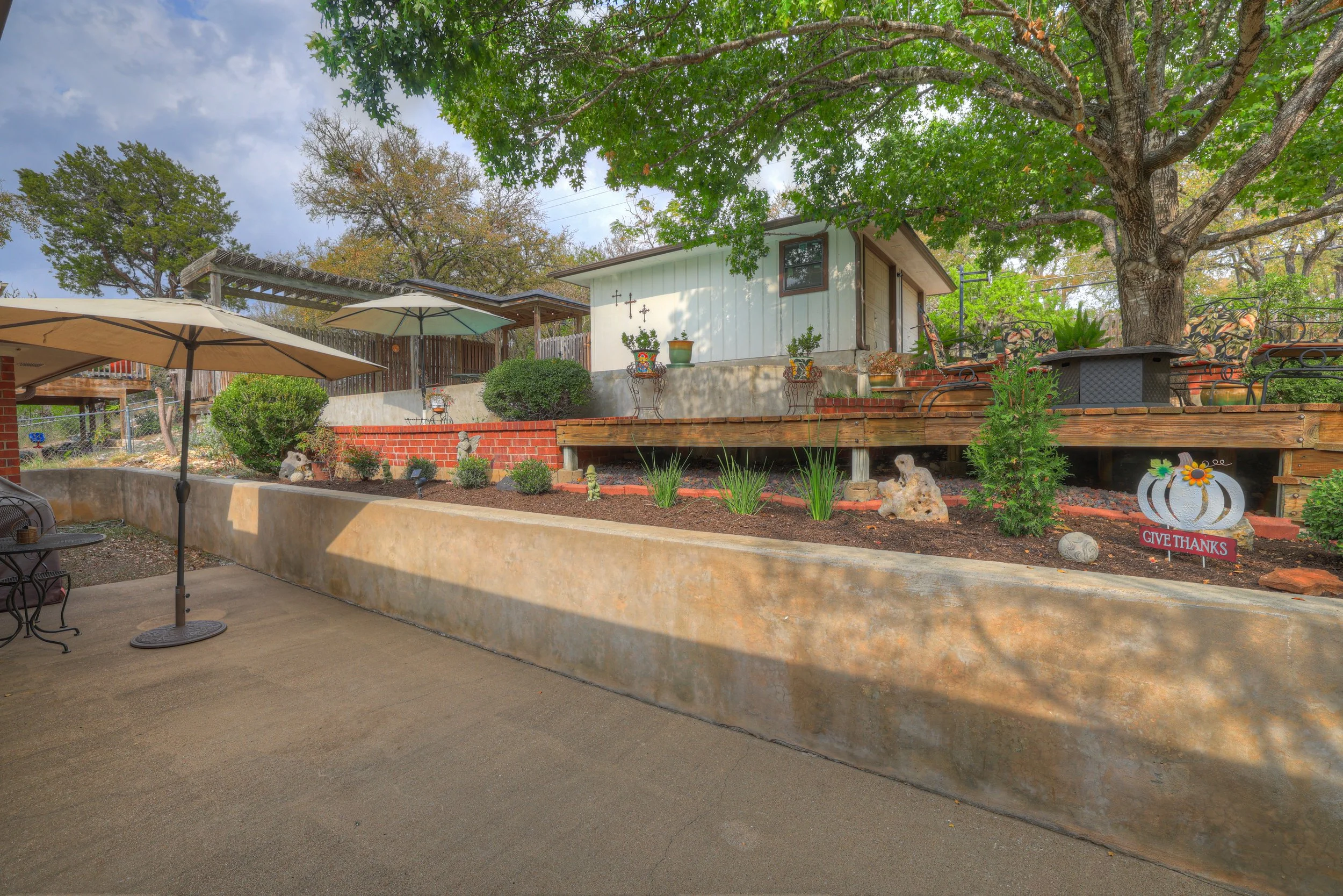 A backyard with a patio area shaded by umbrellas, a raised garden bed with plants, decorative rocks, and a sign that reads 'Give Thanks', with a large tree providing shade, and a small shed in the background.