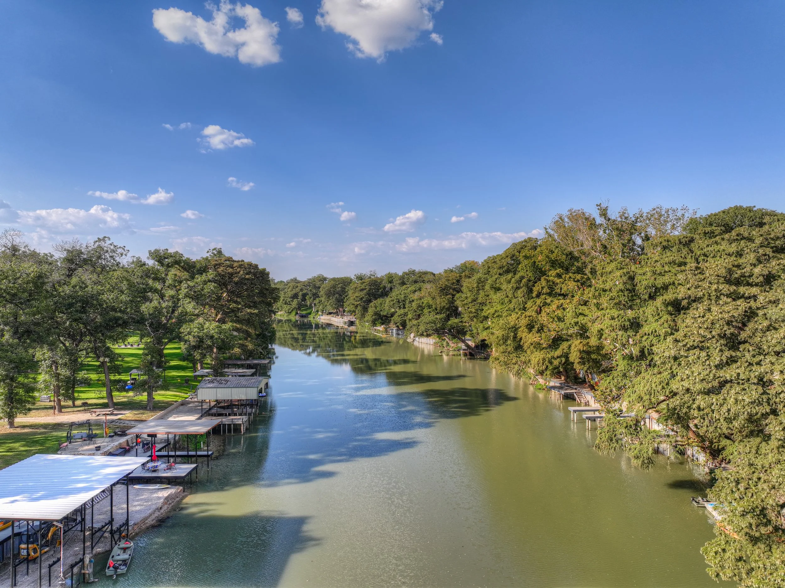 A river lined with trees on both sides, with houses and boat docks along the shoreline under a blue sky with scattered clouds.