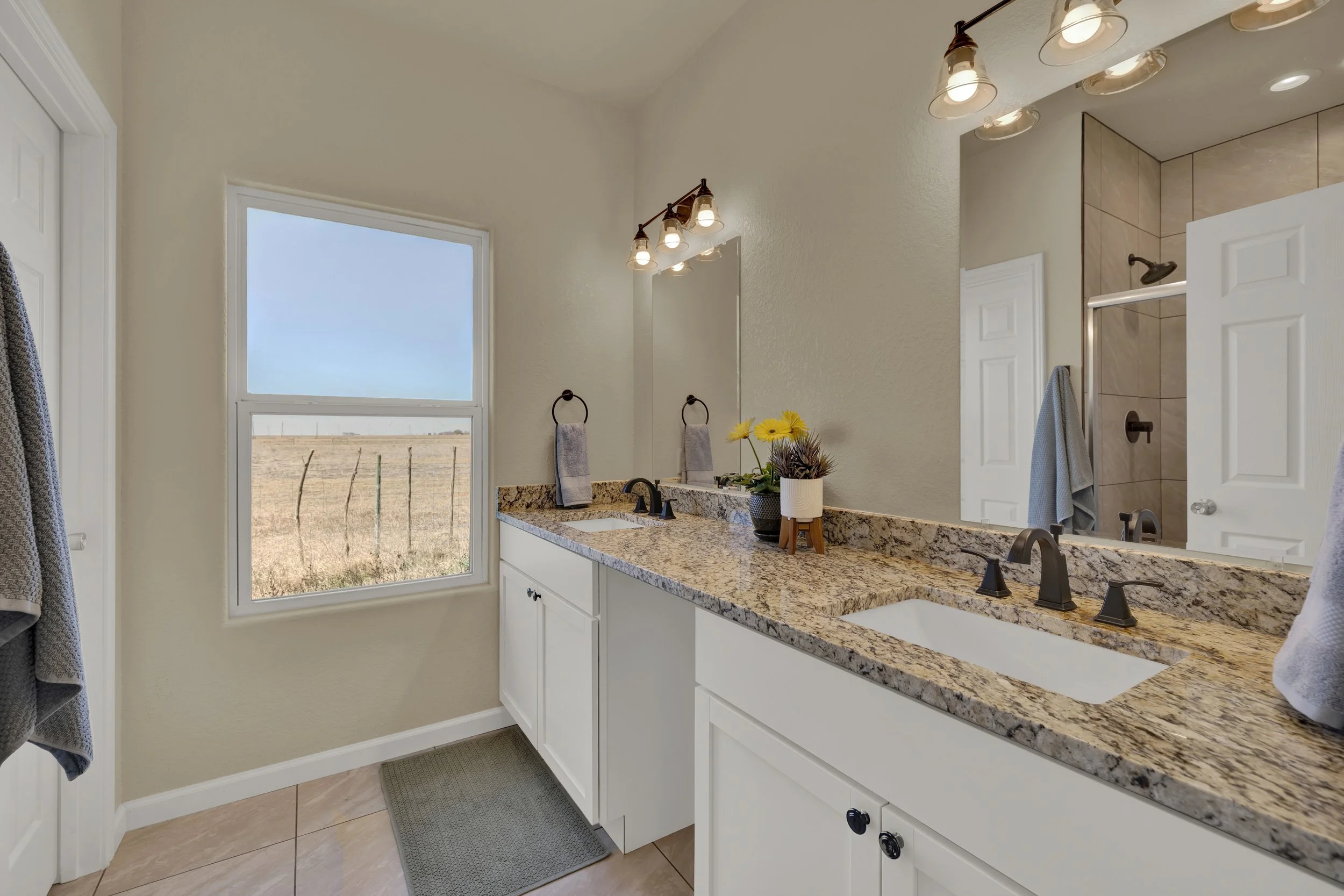 Bathroom with a granite countertop, double sinks, and a large mirror. There are three light fixtures above the mirror; two above the left sink and one above the right sink. The bathroom has beige tile flooring, white cabinets, and a window showing an