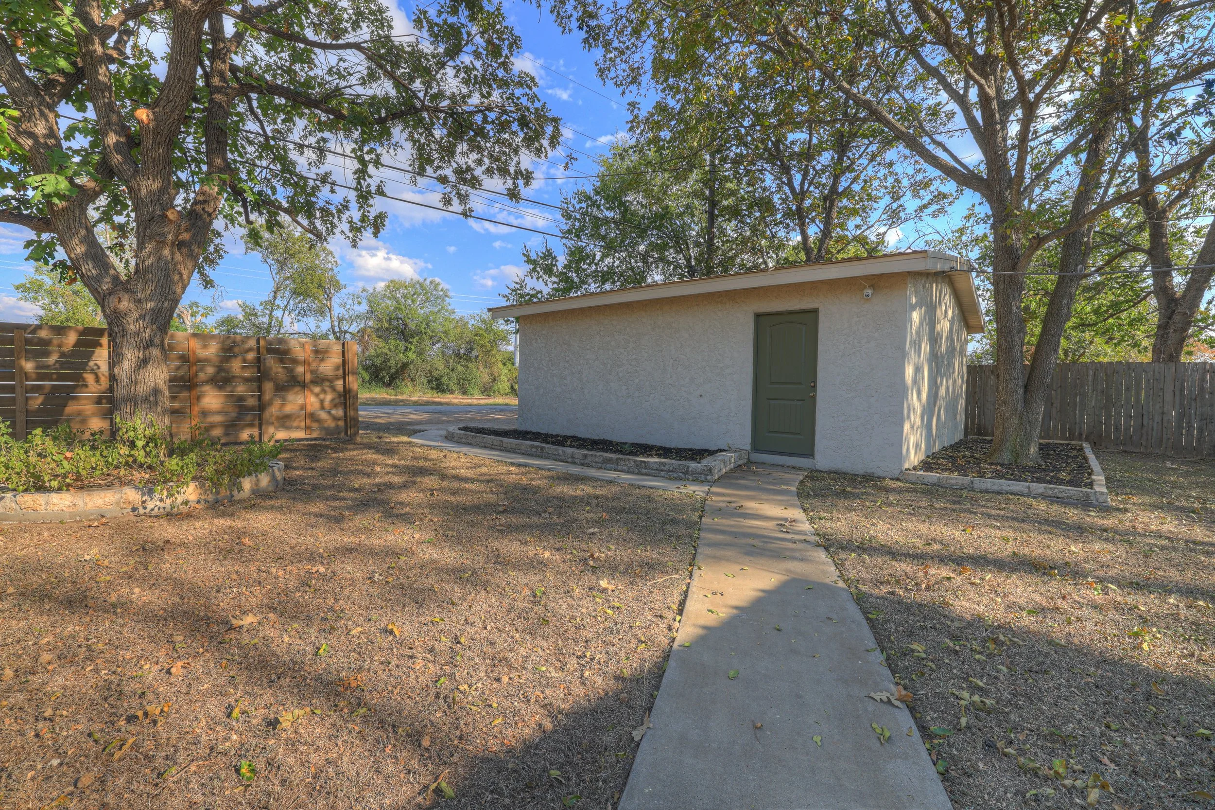 A small white building with a green door, surrounded by two large trees, a wooden fence, and a curved concrete walkway under a blue sky with a few clouds.