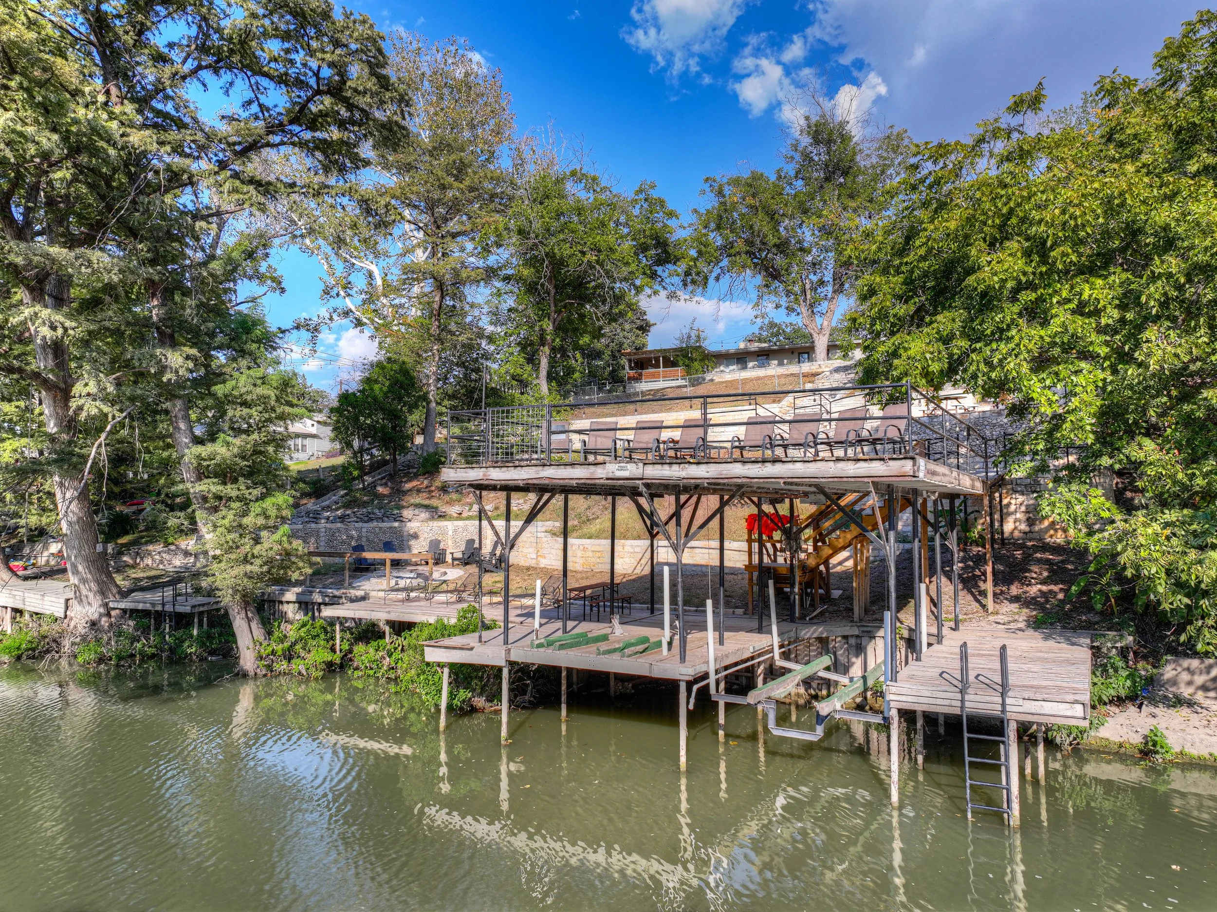 A lakeside view showing a multi-level wooden deck with seating, stairs, and a bar area, surrounded by trees with a blue sky overhead.
