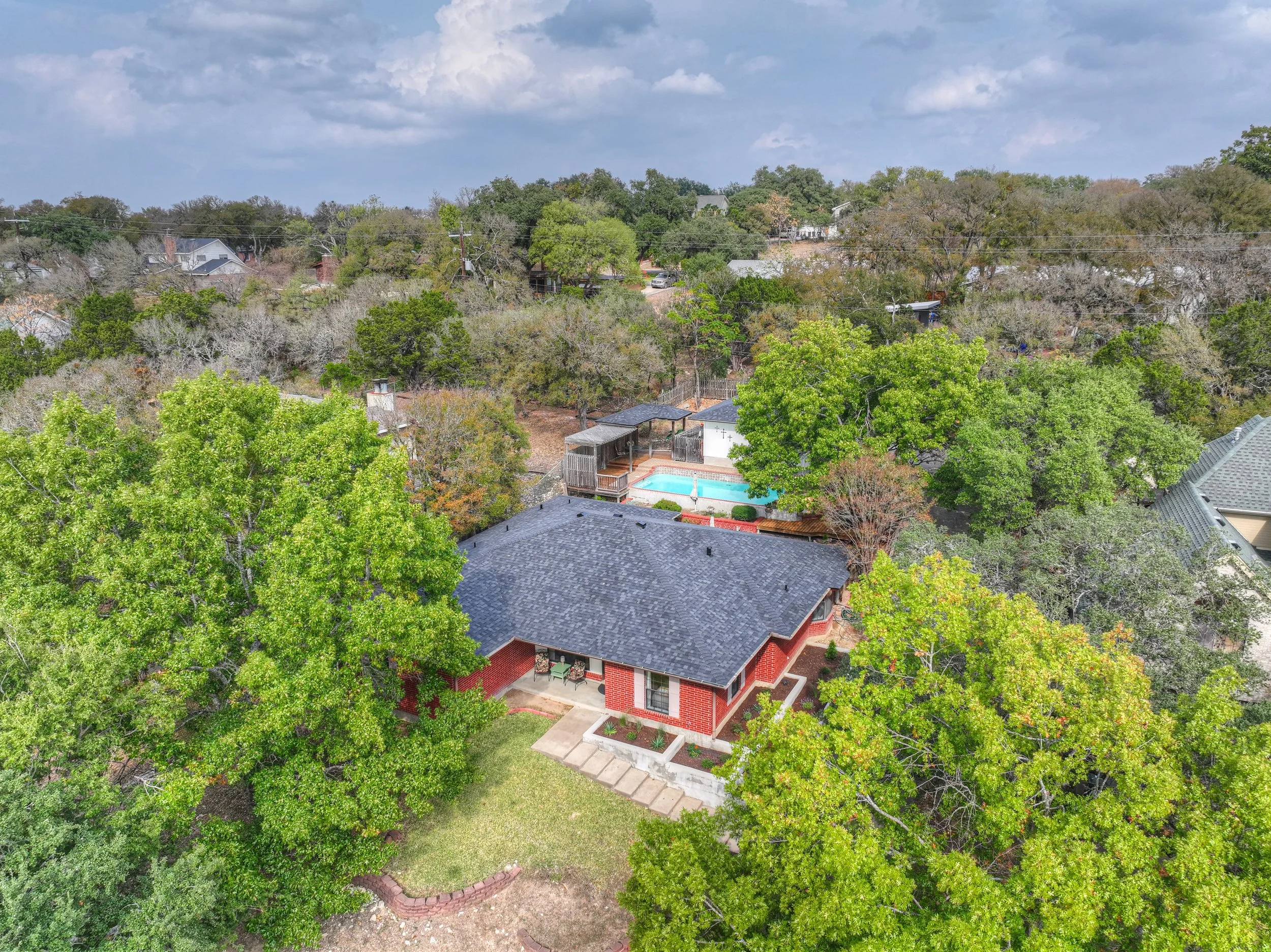 Aerial view of a backyard with a brick house, green trees, a swimming pool with a cover, and a patio area under a partly cloudy sky.