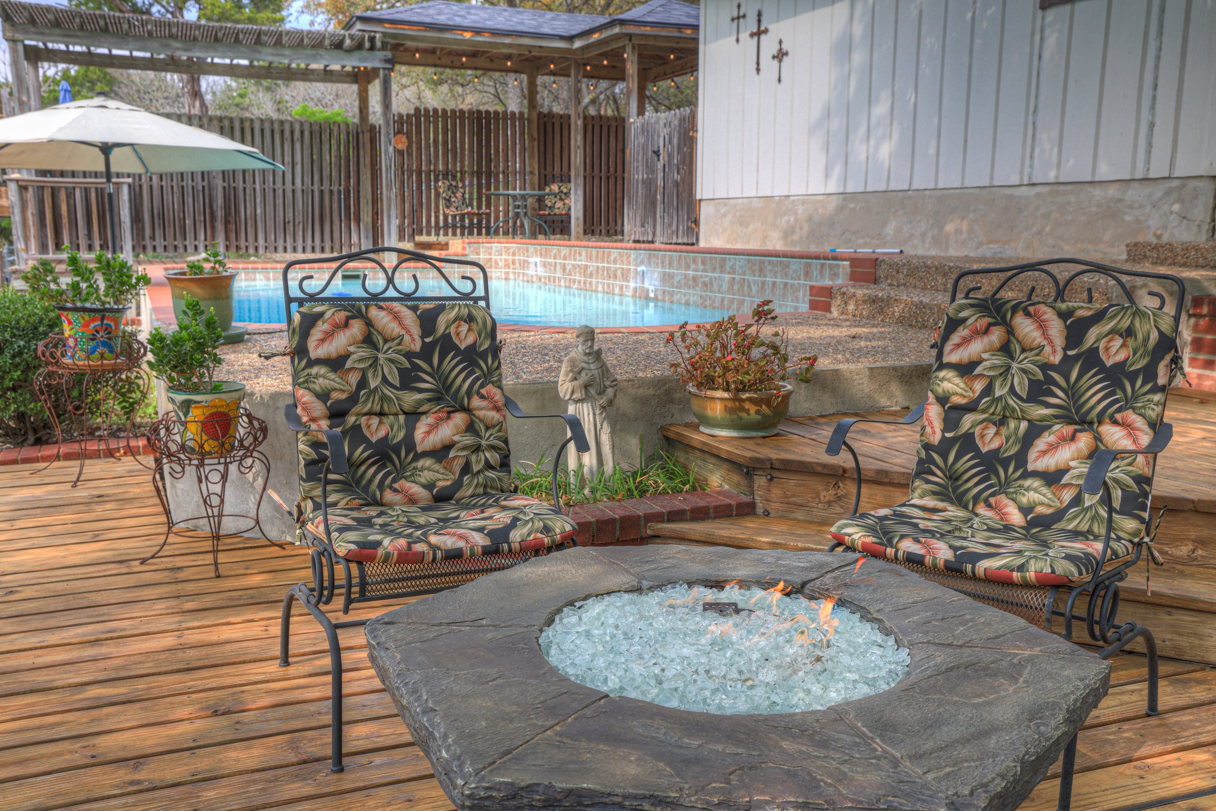 Backyard patio with two chairs featuring floral cushions, a fire pit, potted plants, a swimming pool, and a wooden deck.