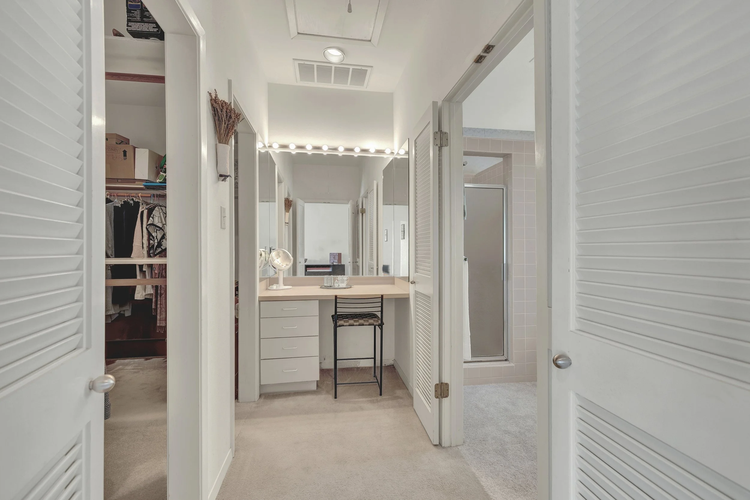A bathroom with a vanity, mirror, and a chair, connected to a walk-in closet and a shower with a glass door.