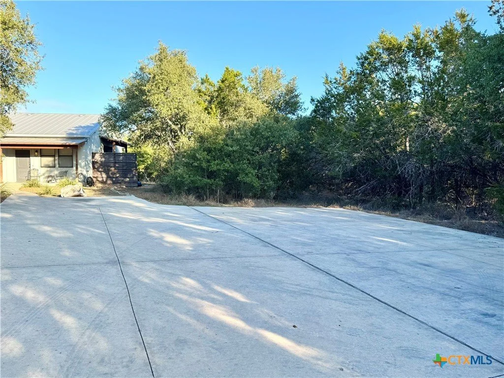 Empty concrete driveway leading to a house with trees and bushes on the sides and a bright blue sky overhead.