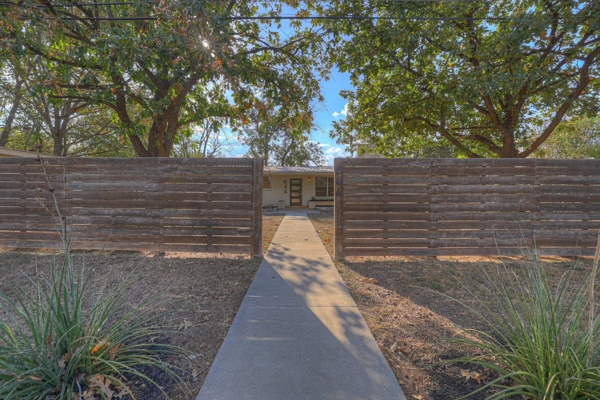 Front pathway leading to a single-story house with a wooden fence on both sides, surrounded by trees with green leaves, under a sunny sky.