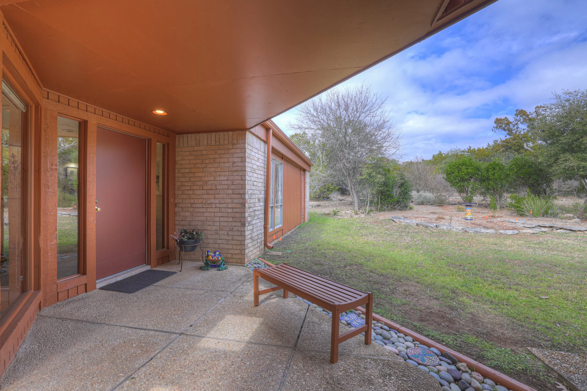 Back porch area of a house with a brown door, a bench, potted plants, and a view of a yard with trees and rocks.