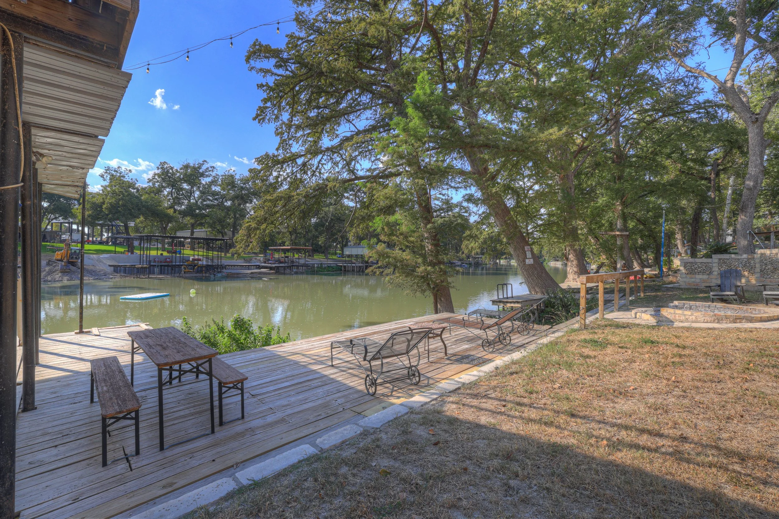 A lakeside scene with a wooden dock and benches, large trees providing shade, and a calm water body with floating paddleboards, under a clear blue sky.