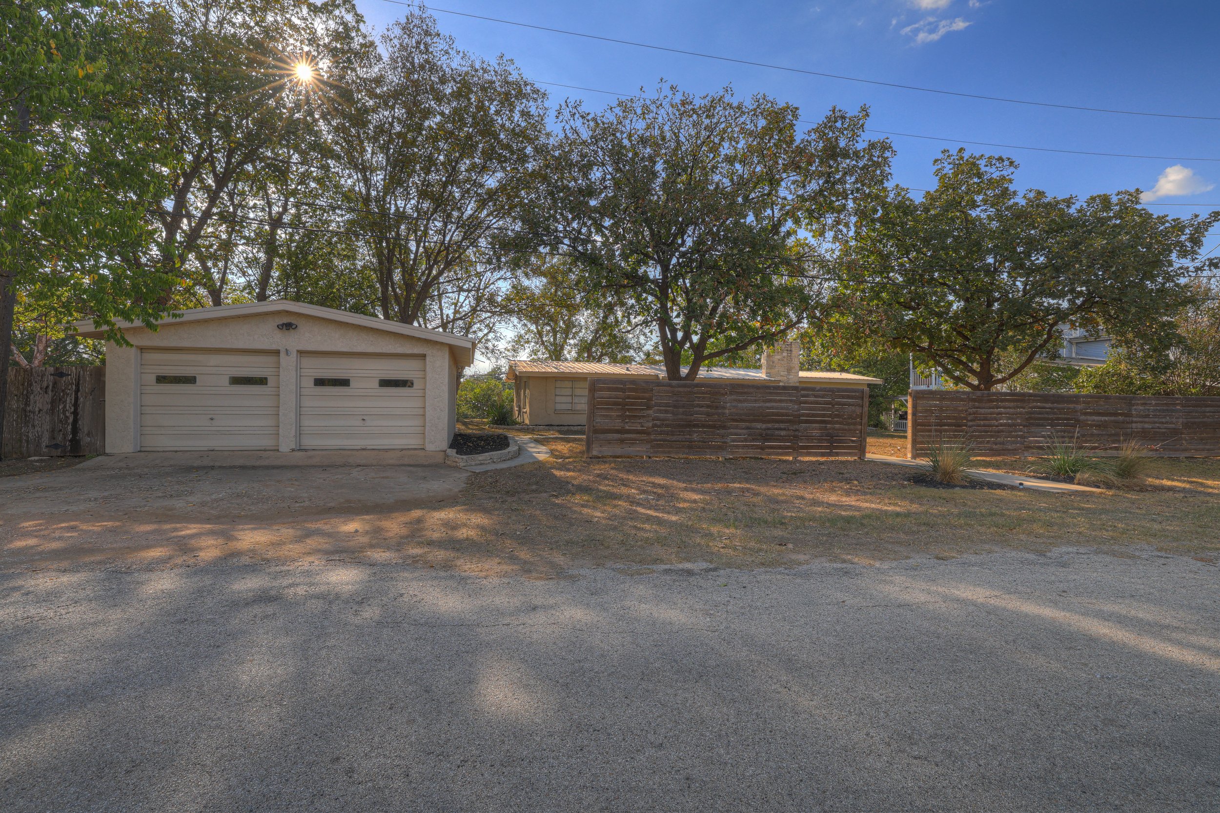 Residential backyard scene with a small house and garage, two large trees, and a wooden fence, under a clear blue sky with sunlight shining through.