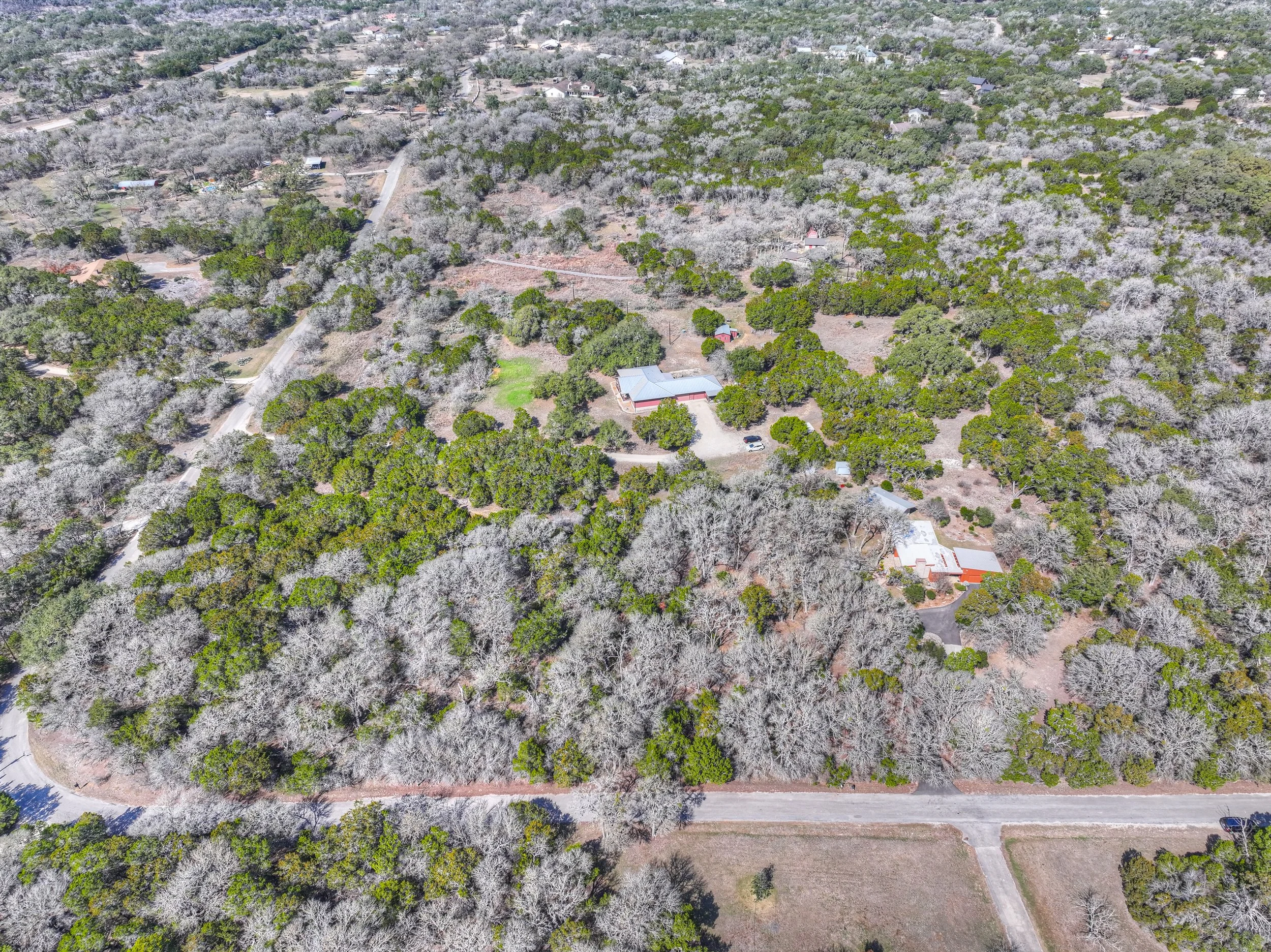 An aerial view of a wooded area with houses, roads, and a mixture of green and leafless trees, suggesting late fall or winter.