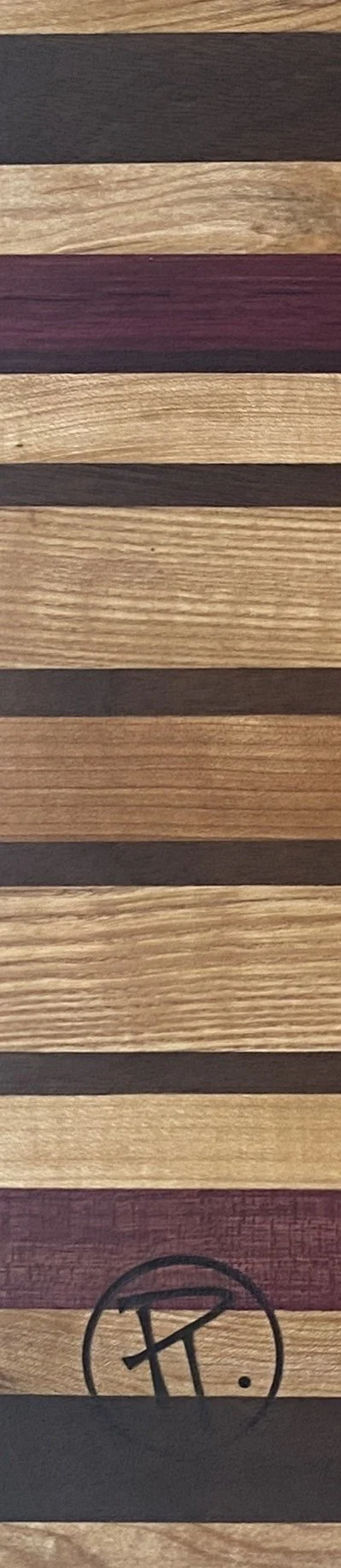Close-up of a wooden surface with alternating dark and light horizontal stripes, featuring a logo with stylized letters and a small dot at the bottom.