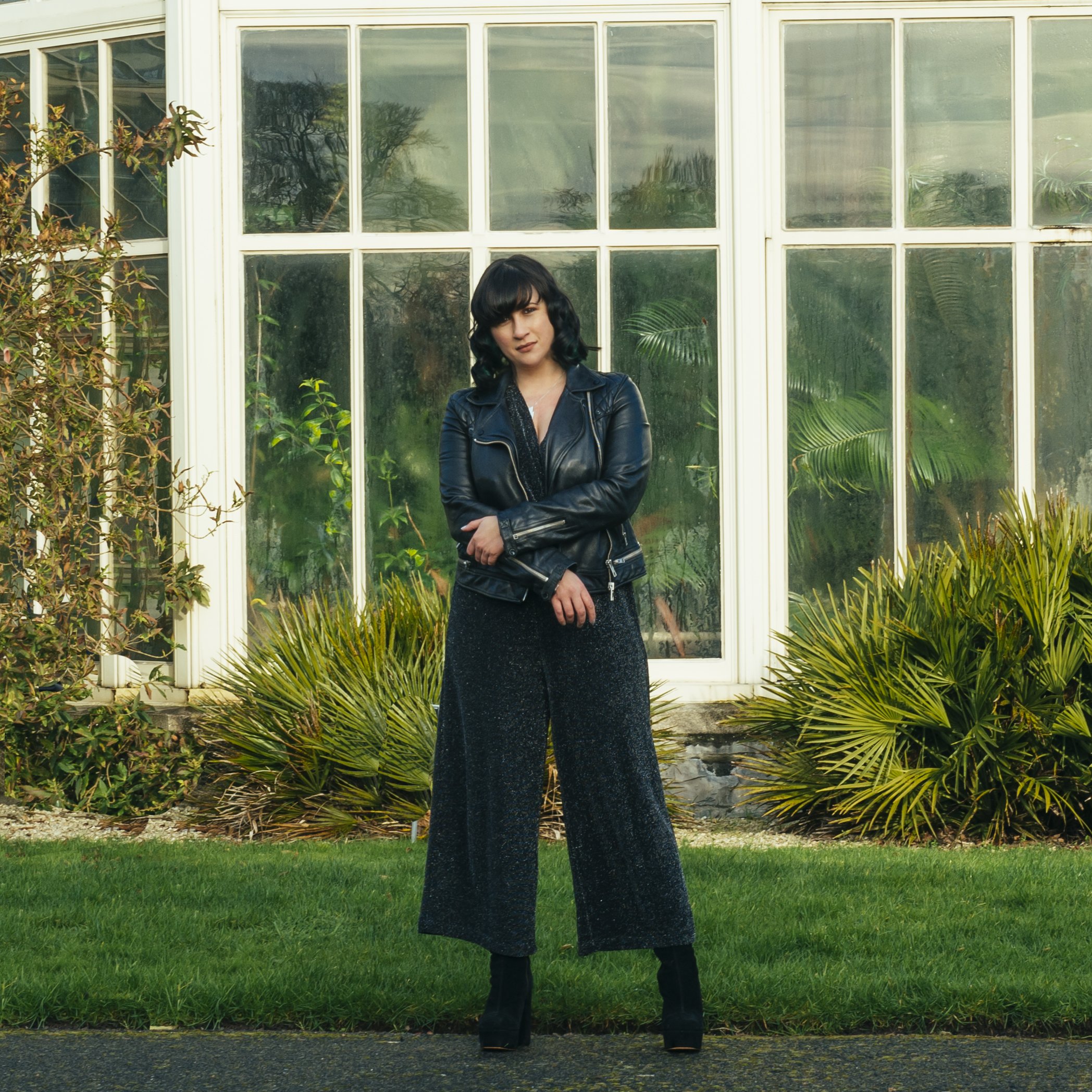 A woman with dark hair in curls standing in front of a greenhouse, wearing a black leather jacket, black sparkly wide-leg pants, and black boots.