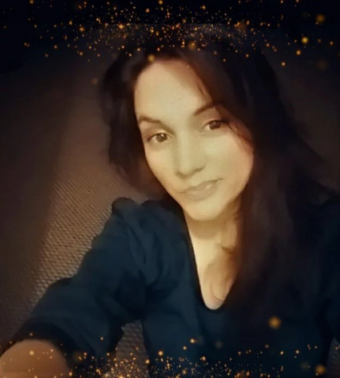 A woman with long dark hair taking a selfie, looking at the camera with a slight smile, against a dark background with a glittery gold overlay at the top and bottom of the image.