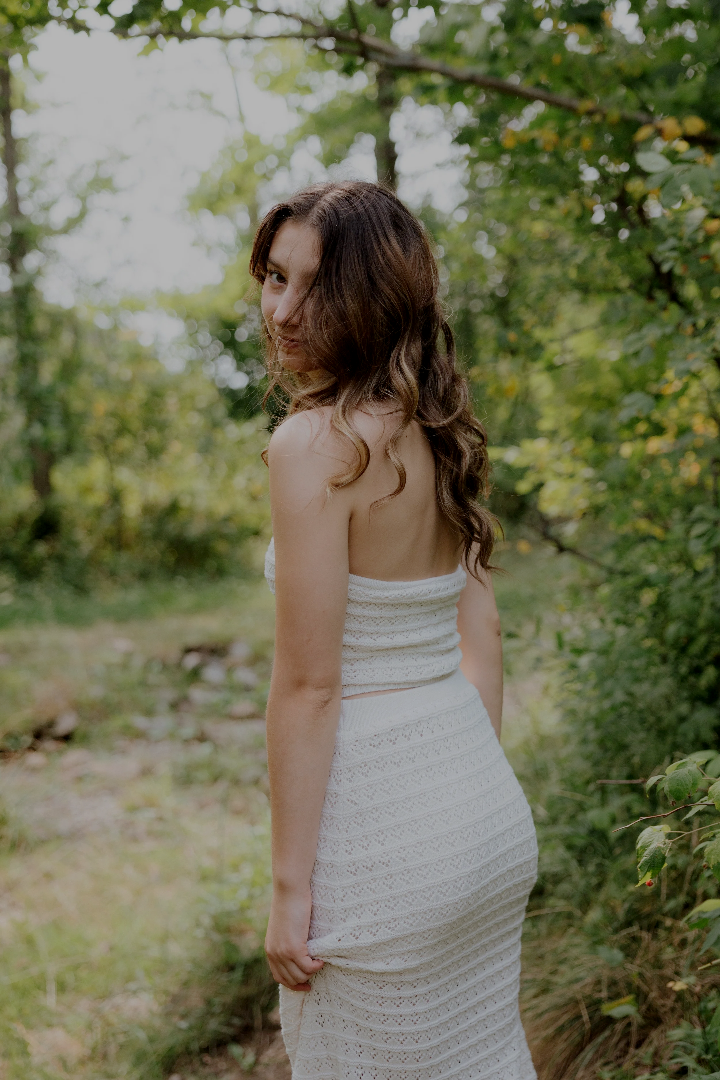 A woman with long, wavy brown hair standing outdoors in a green, wooded area. She is turned slightly away from the camera, smiling and looking over her shoulder. She is wearing a sleeveless white crochet dress.