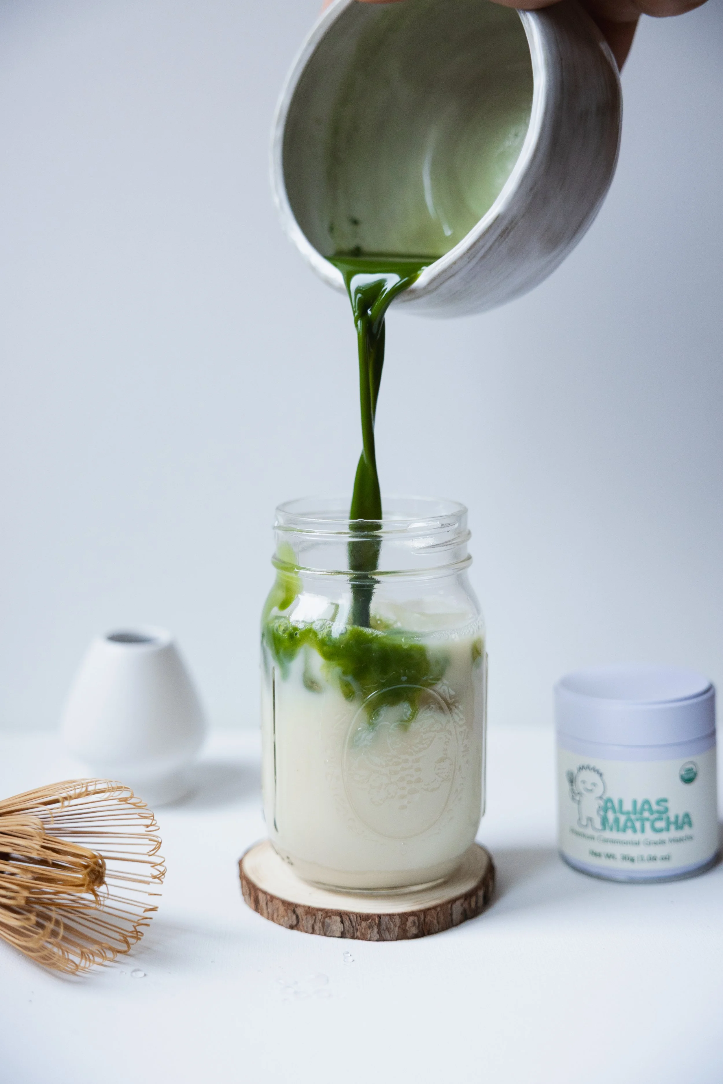 Pouring green matcha latte into a glass mug with a jar of matcha powder and a bamboo whisk nearby.