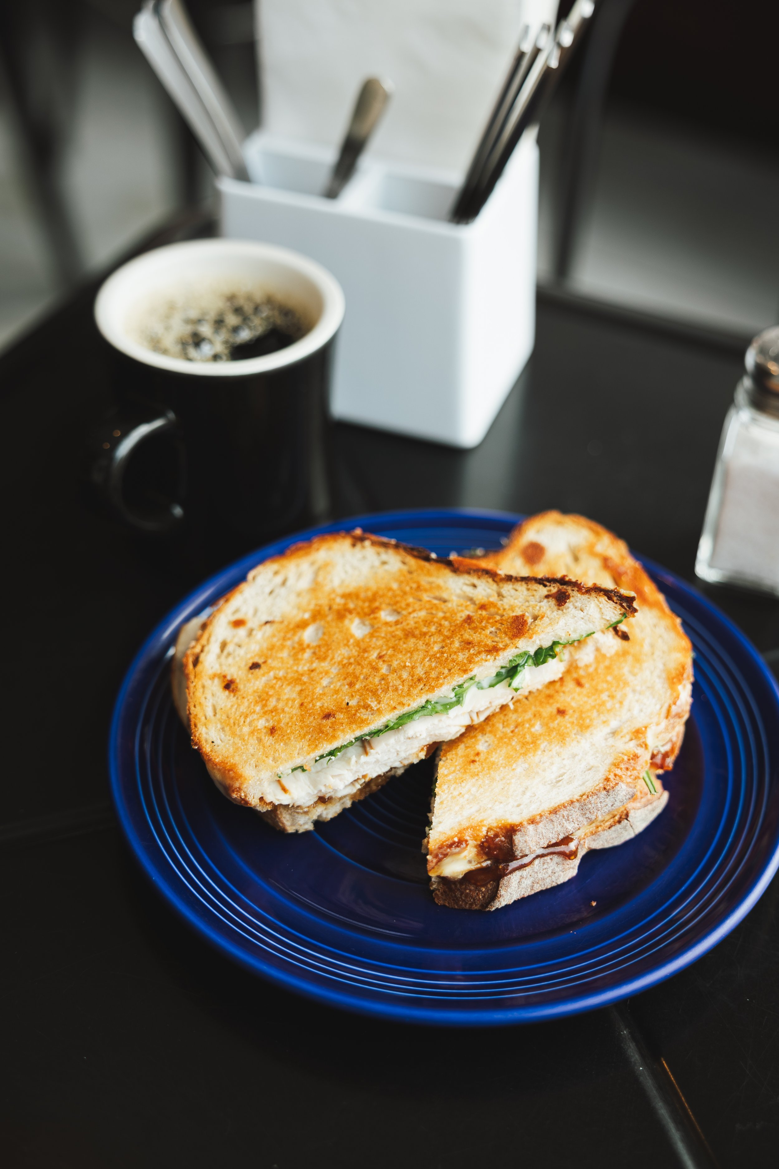 A plate with a grilled sandwich filled with lettuce and chicken, a cup of black coffee, and a container holding utensils on a dark table.