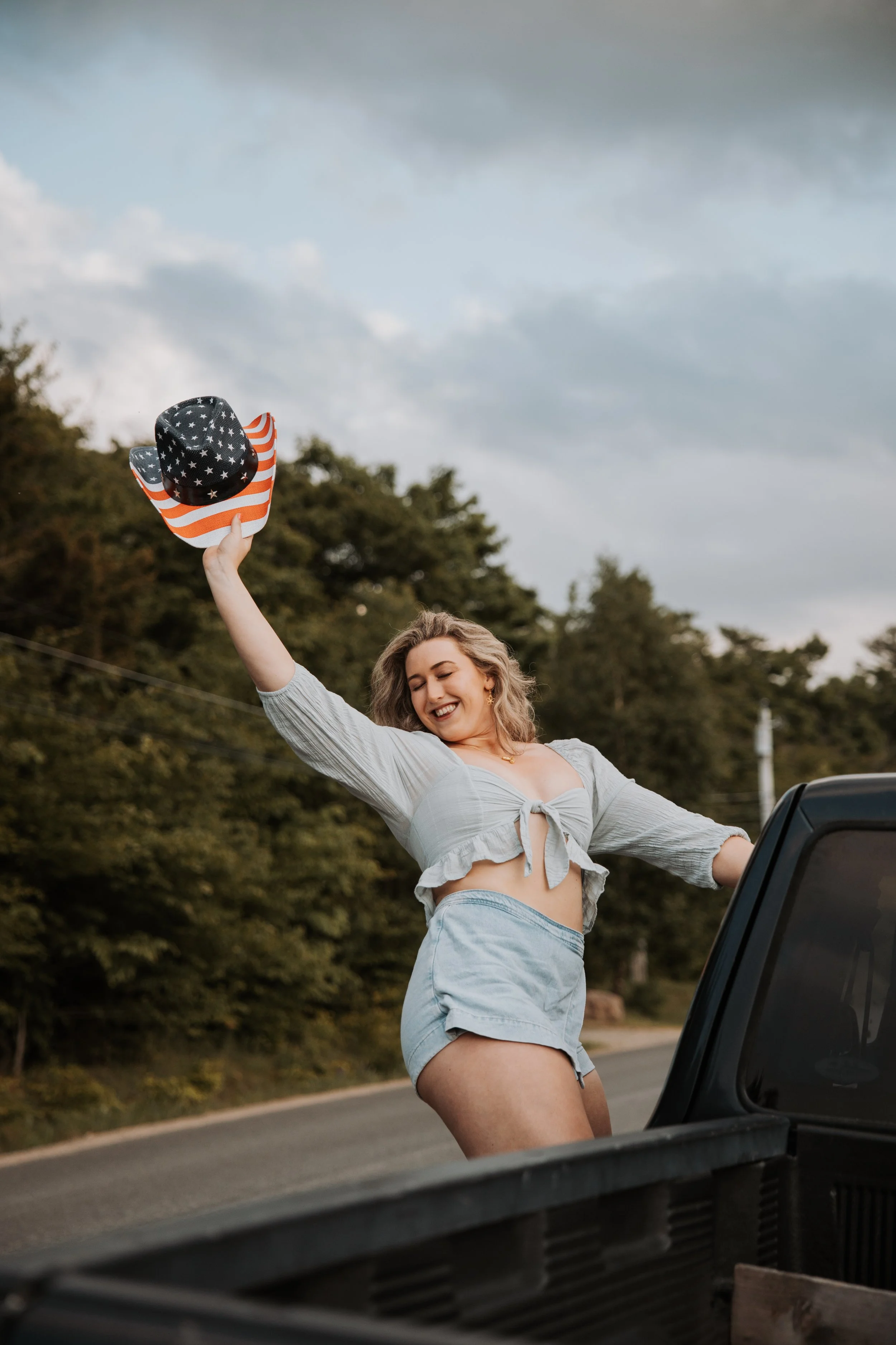 A woman wearing a light gray tied-up crop top and high-waisted shorts is smiling joyfully, holding an American flag-themed cowboy hat up in the air while standing in the back of a pickup truck on a roadside with trees and clouds in the background.